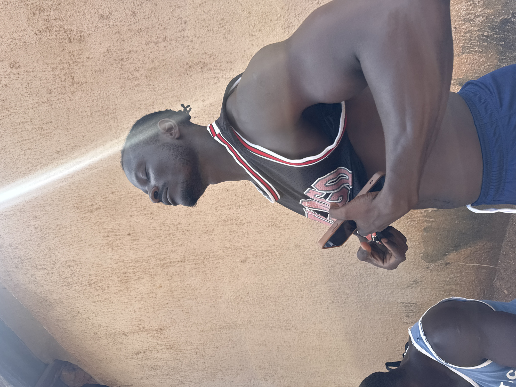 This image, rotated 90 degrees clockwise, presents a close-up of a dark-skinned man, primarily visible from the waist up. He has a beard, short dark hair (possibly dreadlocks or braids), and appears to have his eyes closed. He wears a black basketball jersey with red and white trim, featuring stylized letters that include a prominent "S," indicative of a sports team. He is also wearing blue athletic shorts. In his left hand, he holds a light-colored smartphone with a light brown case, looking down at it. The man exhibits a muscular build.
In the bottom right corner, another dark-skinned person is partially visible, showing their upper back and shoulder, dressed in a light blue or white tank top.
The background is a uniformly textured, light beige or sandy-colored surface, possibly a wall or ceiling. A strong, diagonal beam of sunlight illuminates the upper left portion of the main subject's face and the background, suggesting an outdoor setting during daytime. The ground beneath them appears to be a similar textured surface. The scene is located in Akwanga, Nigeria.
