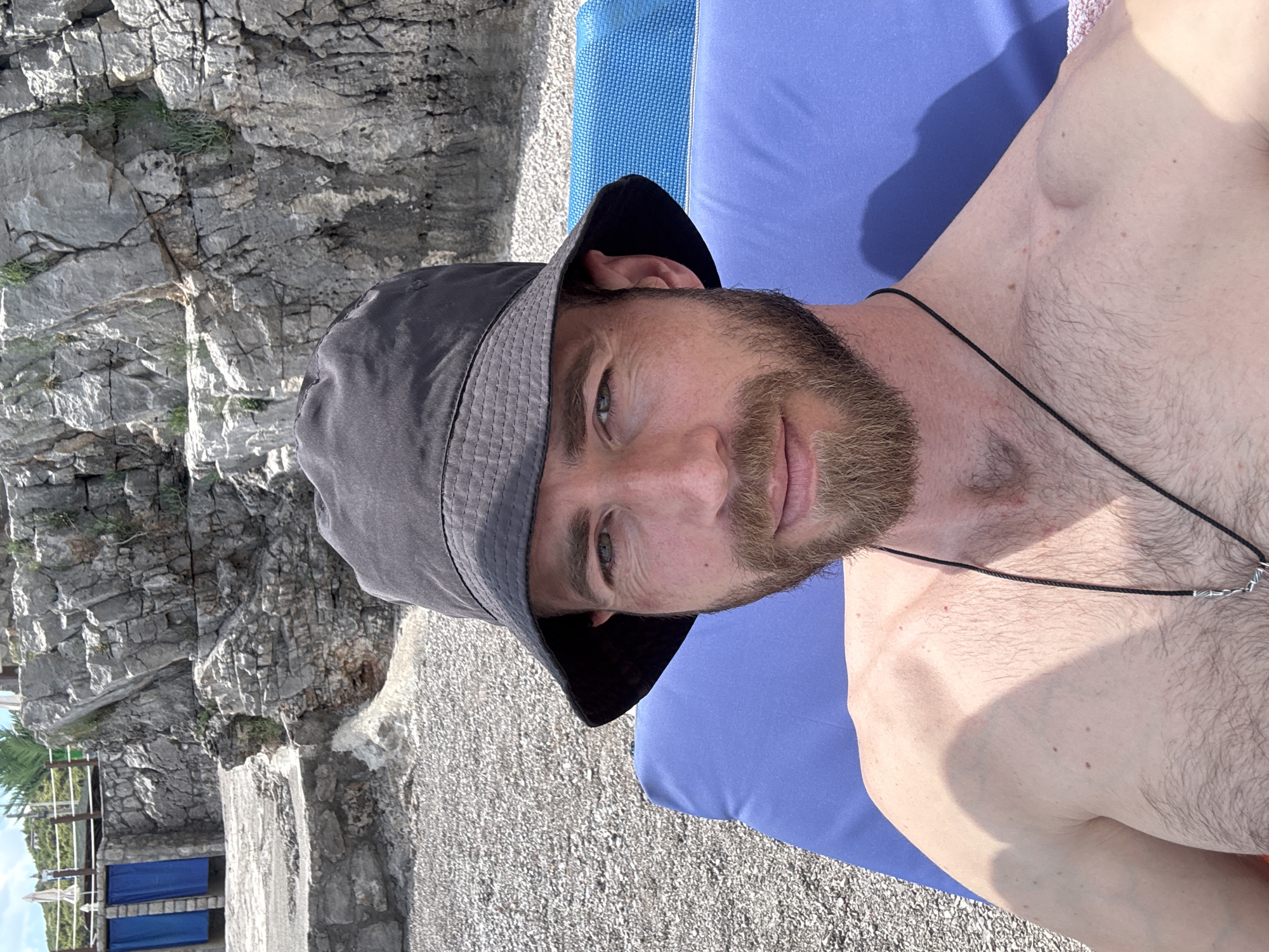 A man is lying on a blue surface on a beach in Prijevor, Montenegro, wearing a gray bucket hat and a necklace. The beach has gravel-like pebbles, and in the background, there is a rocky cliff face. A small structure with blue elements is visible in the lower left corner. The man is fair-skinned, with a beard.