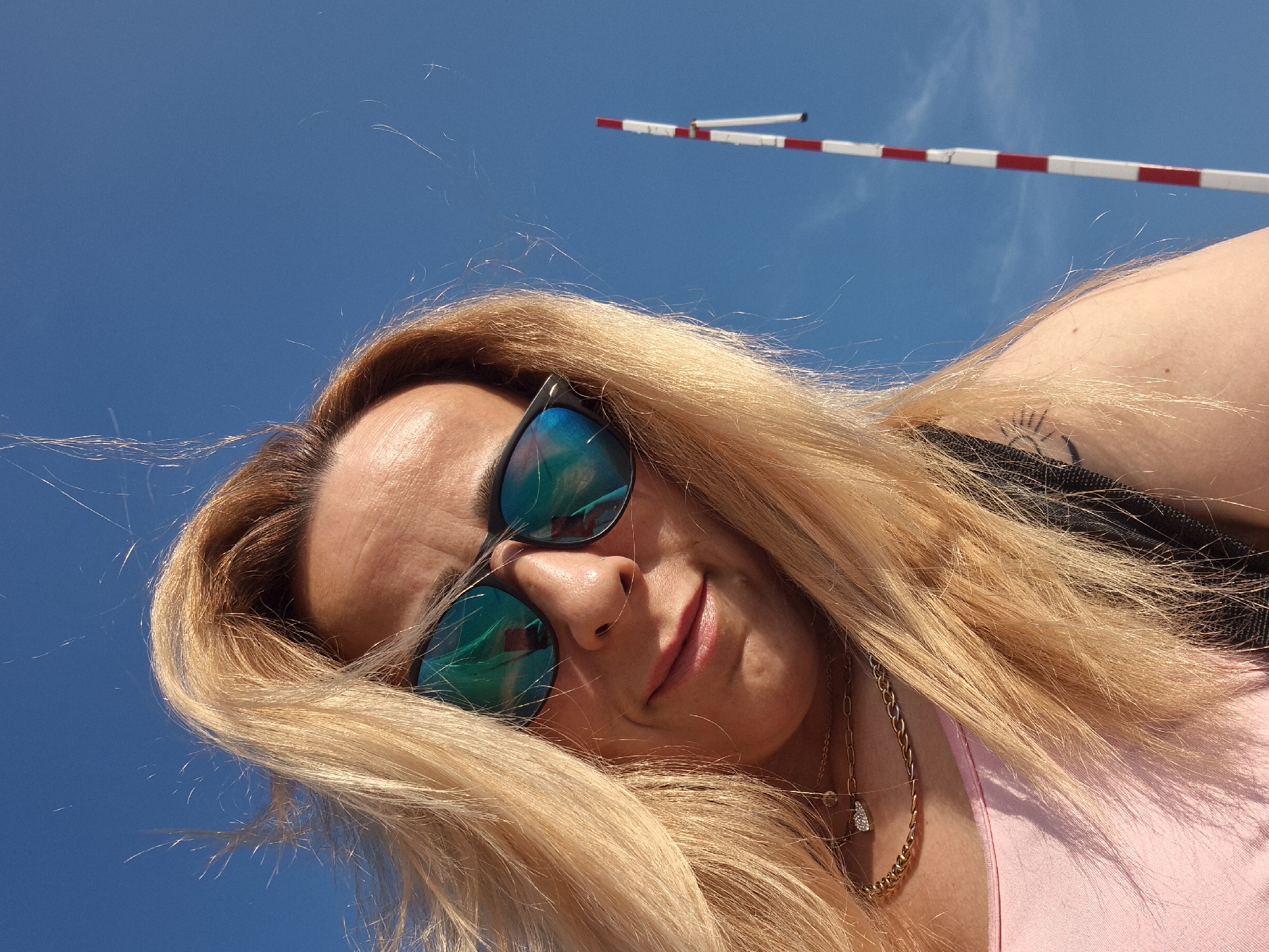 A low-angle, close-up portrait shows a person with long, windswept blonde hair against a clear blue sky. The individual wears dark-framed sunglasses with reflective blue-green lenses, which show reflections of a smartphone held by the photographer. A slight smile is present, and lips are colored pink. Two gold-toned necklaces are visible: a delicate chain with a heart-shaped pendant and a bolder, flat link chain. The person wears a pink tank top or similar garment. A small, dark tattoo is visible on the upper right arm, and a dark shoulder strap, likely from a bag, is visible on the left. On the right side of the background, a tall, slender pole with alternating red and white horizontal stripes extends vertically, featuring a small dark clip or attachment near its top. The scene is brightly lit by natural daylight, indicating an outdoor setting in Unknown, Unknown.