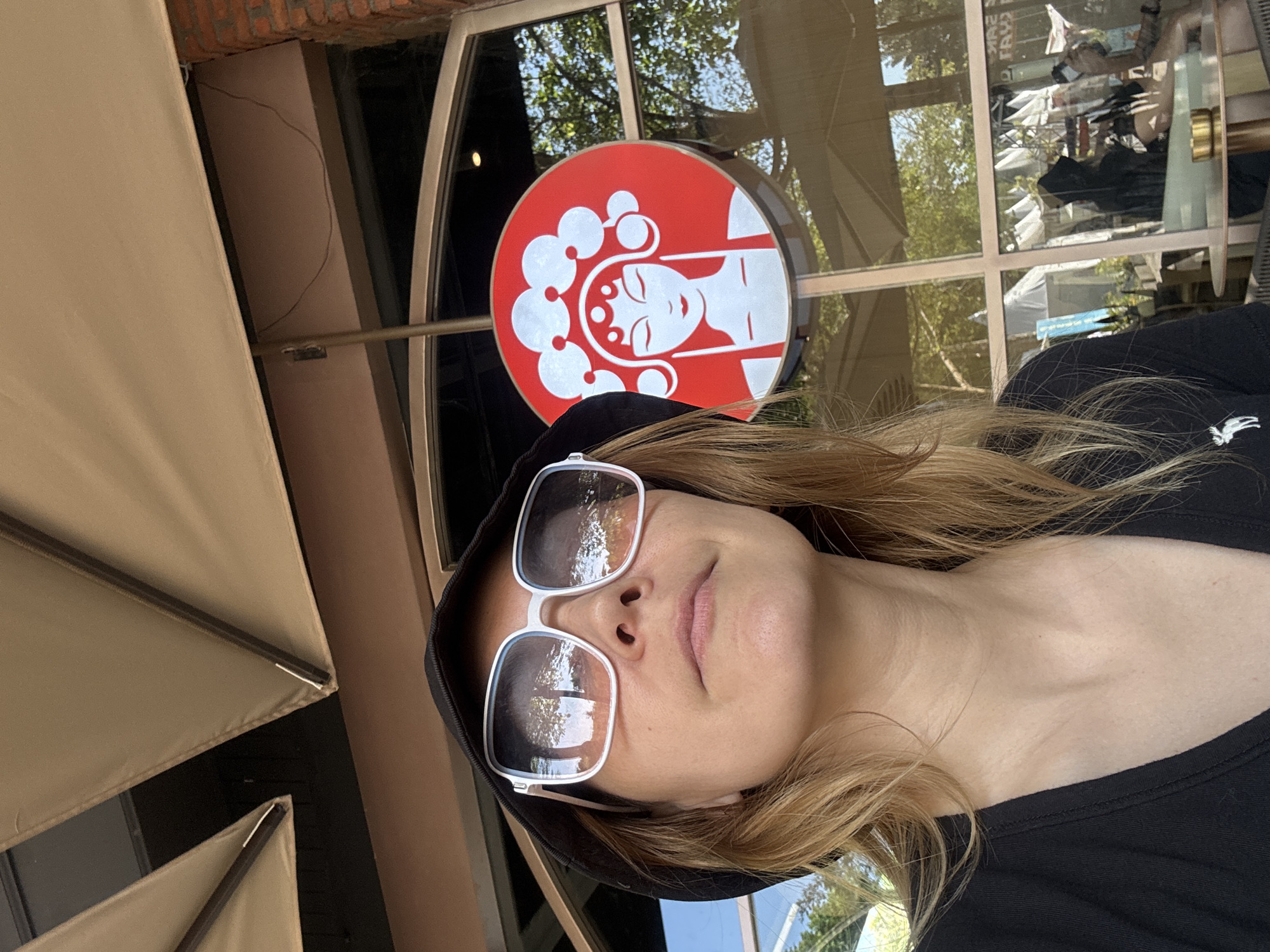 A woman wearing a black bucket hat and large white-rimmed sunglasses is photographed from a low angle, looking upwards and to her right. She has blonde hair and is wearing a black t-shirt with a small white moose logo on the left chest. Behind her, a red circular sign with a white depiction of a woman's face wearing a headdress is visible on a glass window. The background shows an outdoor seating area with tables and chairs, as well as awnings and trees. The image appears to have been taken during the daytime, with sunlight reflecting off the sunglasses and the windows. The location is likely a cafe or restaurant in Kuta Selatan, Indonesia, possibly near the beach, given the reflections of palm trees in the sunglasses.