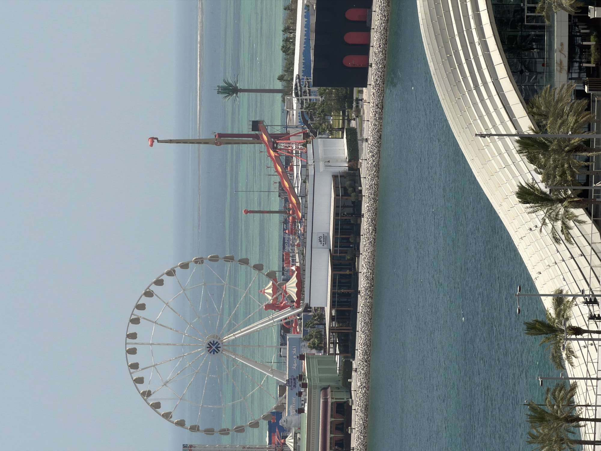 An aerial, wide shot shows a coastal amusement park under a clear, pale sky. The park is situated on the edge of a body of turquoise water. A large white Ferris wheel dominates the left side of the frame. To the right of the Ferris wheel, a red and yellow drop tower ride and a water slide are visible. Various amusement park structures, including striped tents and buildings with green and white facades, are spread across the middle ground. In the foreground, a modern building with a curved, white roofline is partially visible, overlooking the water. Palm trees are scattered throughout the scene. The water stretches to the horizon, where it meets the sky in a soft, hazy line. The overall impression is one of a bright, sunny day at an amusement park.
