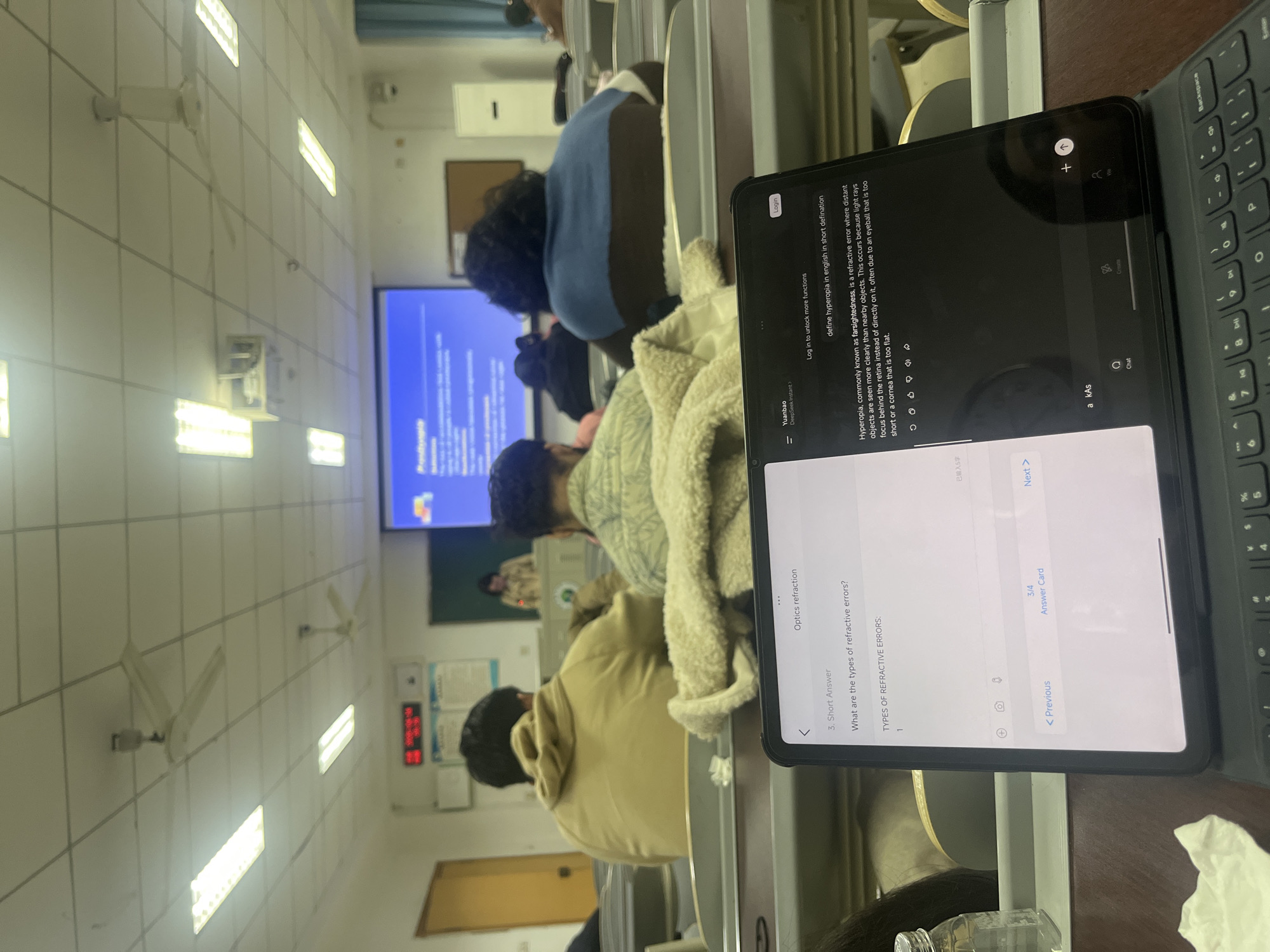 The image depicts a classroom setting with students attending a lecture. A tablet with a keyboard is prominently positioned in the foreground, displaying text related to "hyperopia" and "refractive errors," suggesting the lecture is related to optics. The classroom is brightly lit with fluorescent ceiling lights, and a projection screen shows educational content. In the background, other students are seen seated at desks, listening to the lecture. The exact location within Yuzhong District, China cannot be determined from the image.
