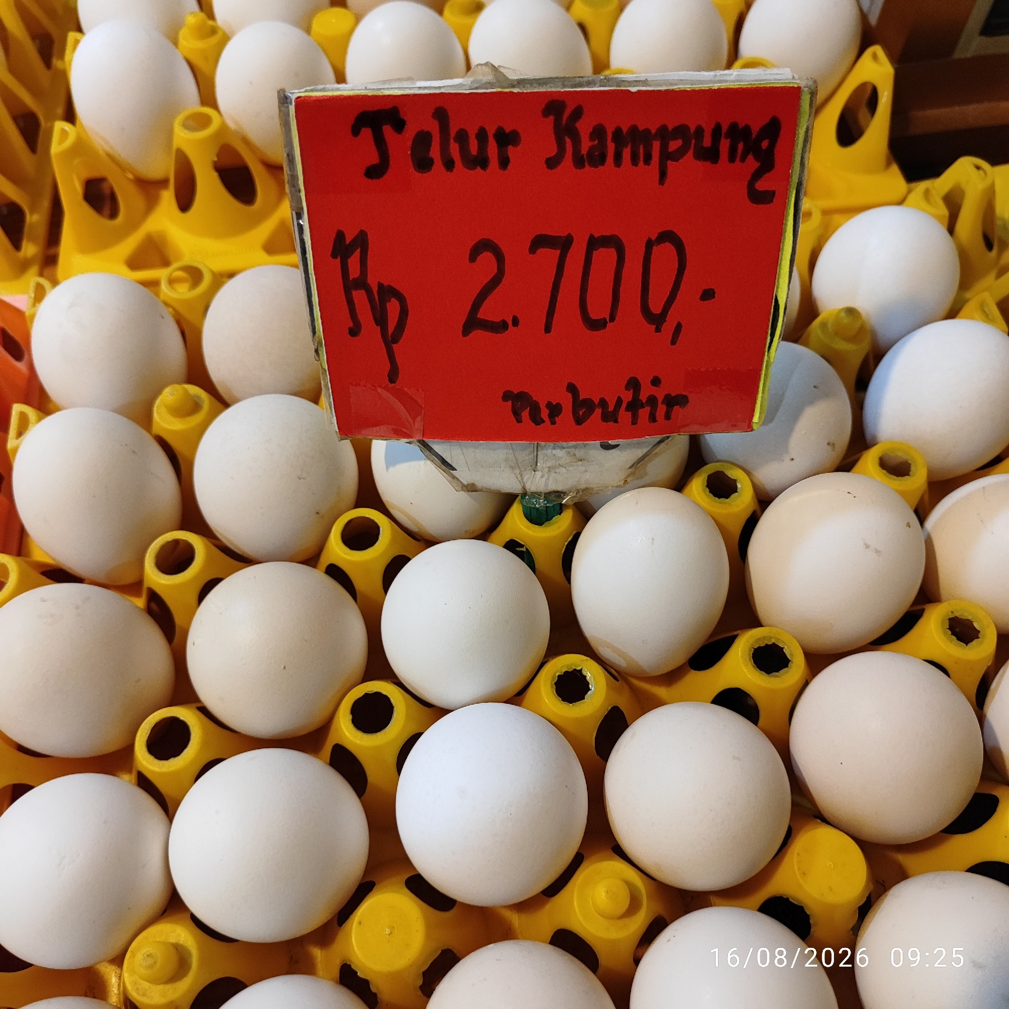 A close-up, overhead view showcases a tray of white eggs. A bright red sign with handwritten text is placed prominently in the center of the tray, partially obscuring some of the eggs. The sign reads "Telur Kampung Rp 2.700,- per butir". The eggs are nestled in yellow plastic dividers, with some eggs visible in the background. The lighting is natural and appears to be from an overhead source. A timestamp, "16/08/2026 09:25", is visible in the bottom right corner of the image. The overall scene suggests a market stall or a grocery store display. The city of Pembaharuan, Indonesia is not discernable from the image content.