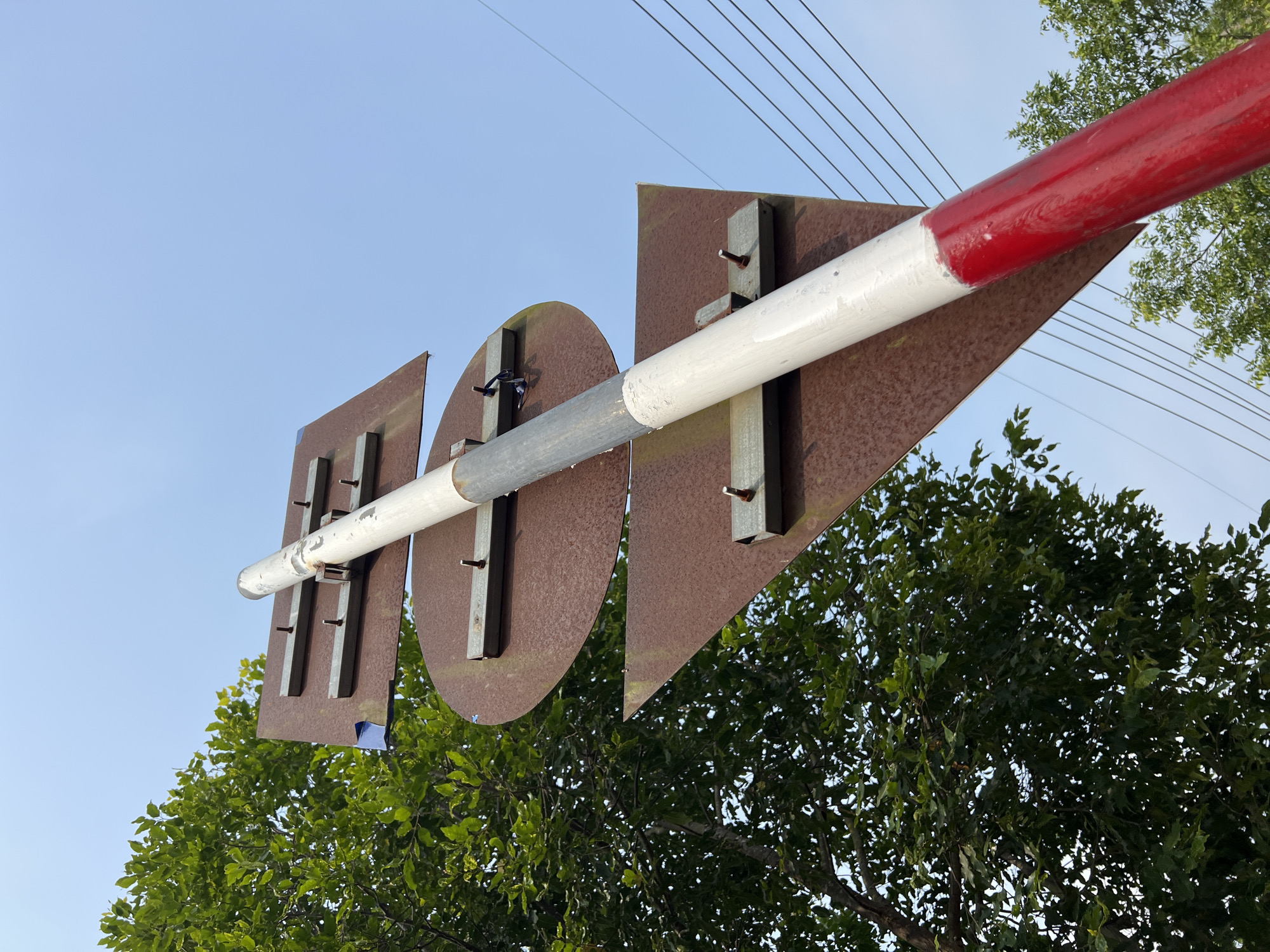 A low-angle shot depicts a red and white striped boom barrier, likely used to control access at a road or railway crossing. The barrier is supported by rusty metal plates shaped like a rectangle, circle, and triangle. The attachments are secured with metal bolts. The background shows a clear blue sky and a dense green tree with visible power lines above. The precise location within Bo Y, Vietnam is not discernible from the image.
