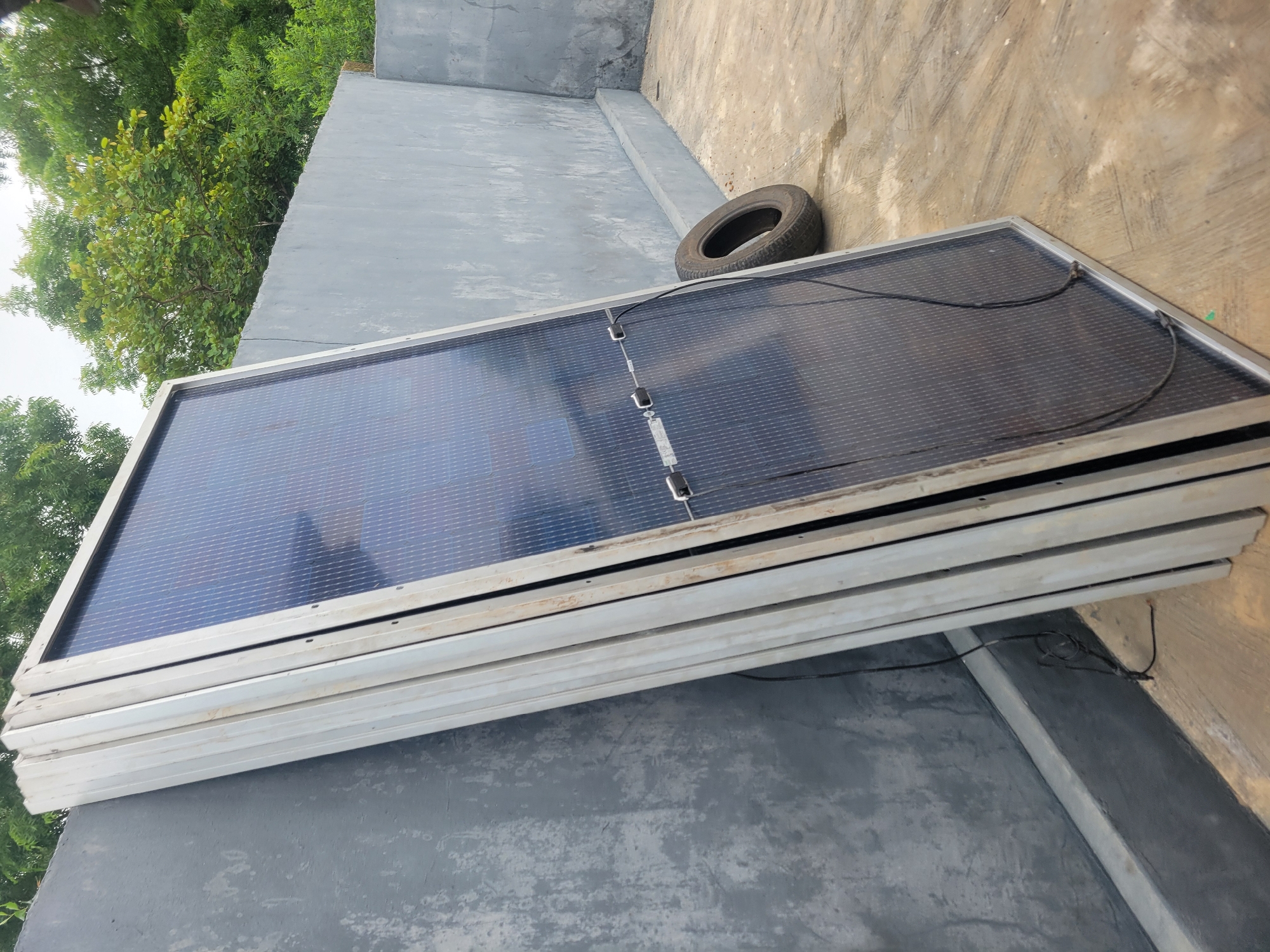 A stack of solar panels leans against a grey, weathered concrete wall outdoors. The panels are silver-framed and have a dark, grid-like surface. A black tire lies on the ground to the right of the panels, near the wall. The ground is a light brown, concrete surface with some debris. In the background, lush green trees are visible above the wall. The lighting suggests it is daytime.