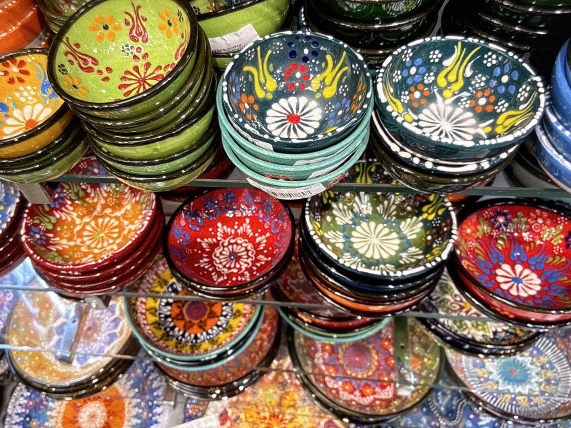 The image shows a close-up, high-angle, full shot of a stack of vibrantly colored, intricately decorated ceramic bowls and plates. The items are arranged vertically and slightly angled, filling the frame. The surfaces of the ceramics display a variety of patterns, including floral motifs, geometric designs, and dots. Colors range from bright reds, oranges, and yellows to greens, blues, and white, often set against darker backgrounds. Some of the items appear to have a glossy finish. The lighting suggests an indoor setting, possibly a market or shop, with some reflections visible on the surfaces. The background is a blur of more stacked ceramics, indicating a large display. The specific location in the city of Orihuela, Spain, is not identifiable from the image content.