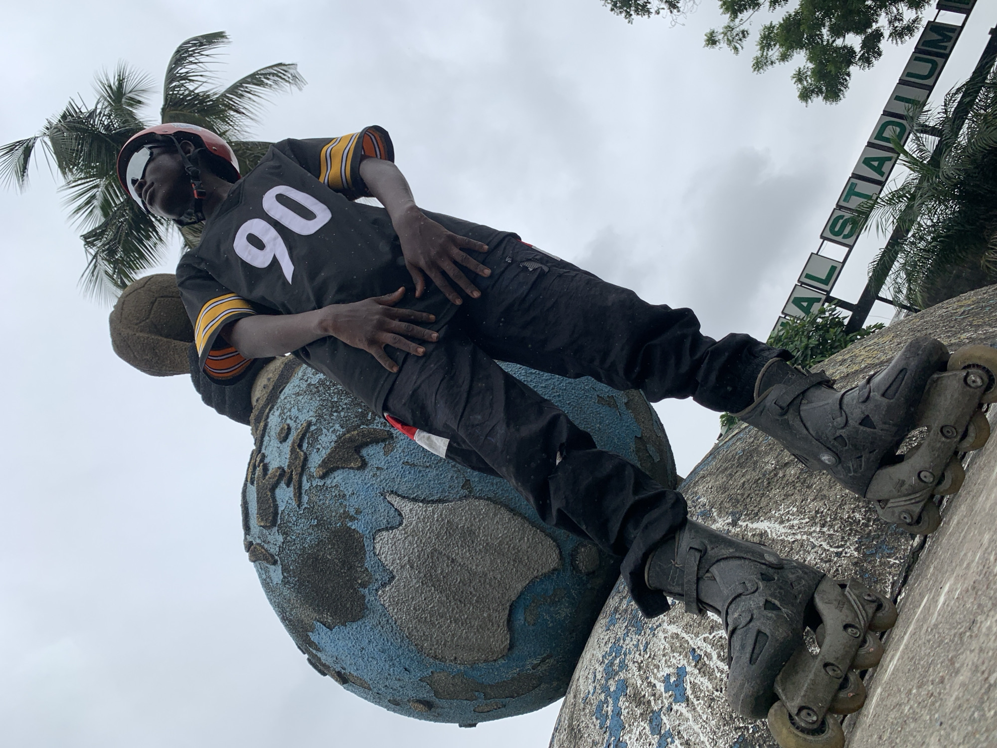 A low-angle, full-body shot captures a young Black male on roller skates, positioned in front of a large, weathered globe sculpture. The man is wearing a black t-shirt with the number "90" in white, a red helmet, and dark cargo pants. He is looking upwards with his eyes closed, and his hands are placed in front of him. The globe sculpture, with visible continents, appears to be part of an outdoor monument, likely at a recreational area. In the background, a palm tree and a sign that reads "AL STADIUM" are visible. The sky is overcast, suggesting a cloudy day. The scene is outdoors, in the city of Iganmu, Nigeria.