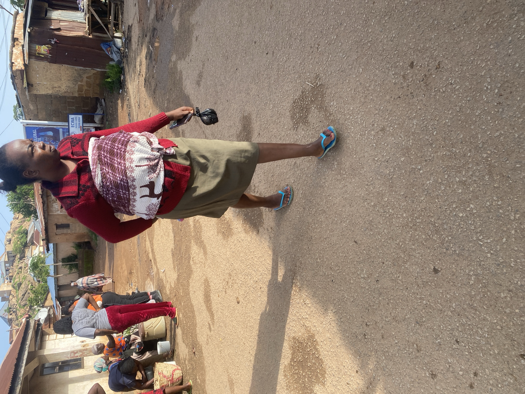 In Jos, Nigeria, a woman with dark hair, wearing a red cardigan and a patterned cloth tied around her midsection, walks on a dirt road. She is wearing a khaki skirt and blue flip-flops. She carries a black plastic bag in her right hand. In the background, other people are seen near buildings and baskets. The area appears to be a residential or market district with a hilly landscape in the distance.