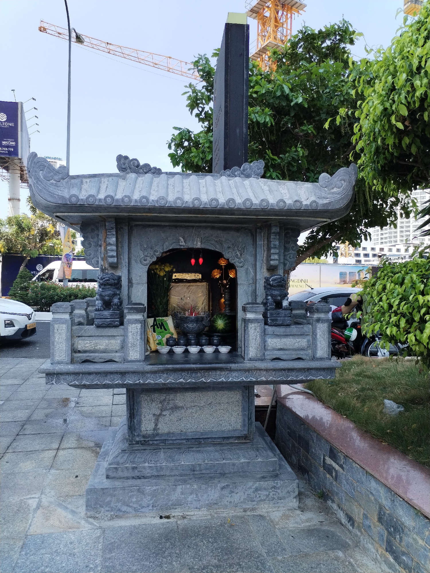 The image captures a small, ornate, stone spirit house on a city sidewalk in Nha Trang Ward, Vietnam. The spirit house has a tiled roof with decorative edges and a small interior space holding offerings: small bowls, a black pot, flowers, and red hanging lamps. The spirit house is located next to a curb with a retaining wall and a patch of grass. In the background are cars, a motorcycle, and a crane. A billboard on the left side of the frame, along with the urban setting, suggests the spirit house's proximity to a road in the ward.