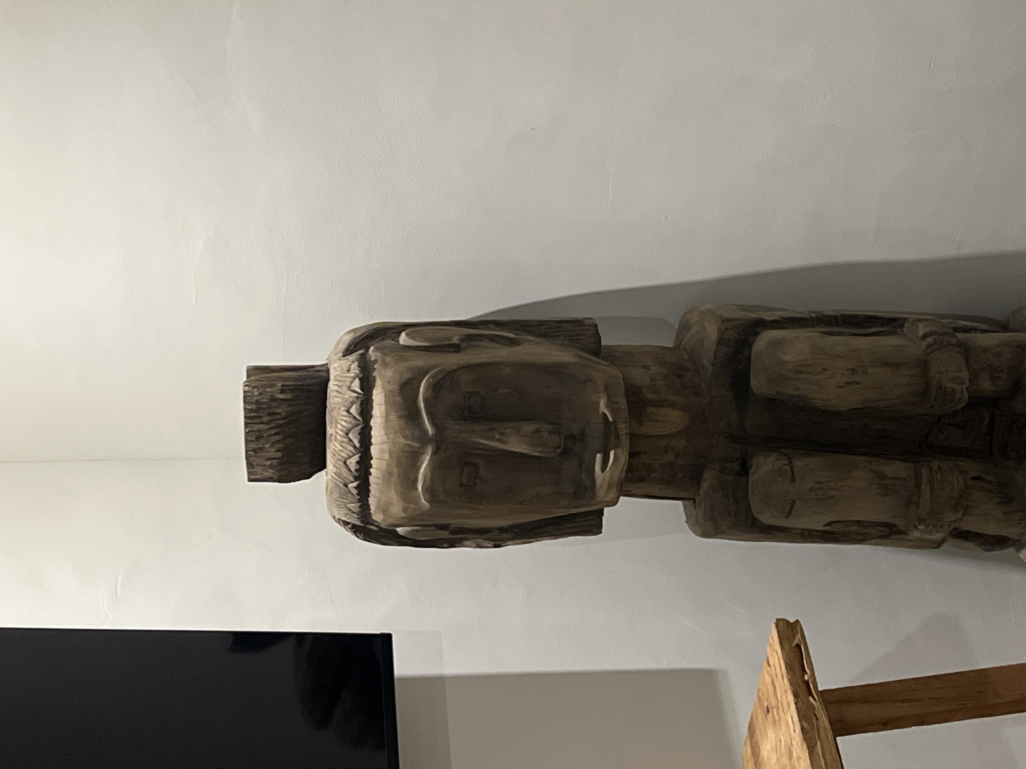 In a room with plain white walls, a carved wooden statue stands against the wall. The statue depicts a human figure with a broad, flat face, prominent nose, and what appears to be a headdress or hairstyle. The body of the statue is carved to suggest folded limbs. To the left of the statue, a portion of a black rectangular object, likely a television screen, is visible. In the lower left of the frame, the edge of a wooden table with a rough-hewn surface and visible grain is present. The lighting in the room casts a shadow of the statue onto the wall behind it.