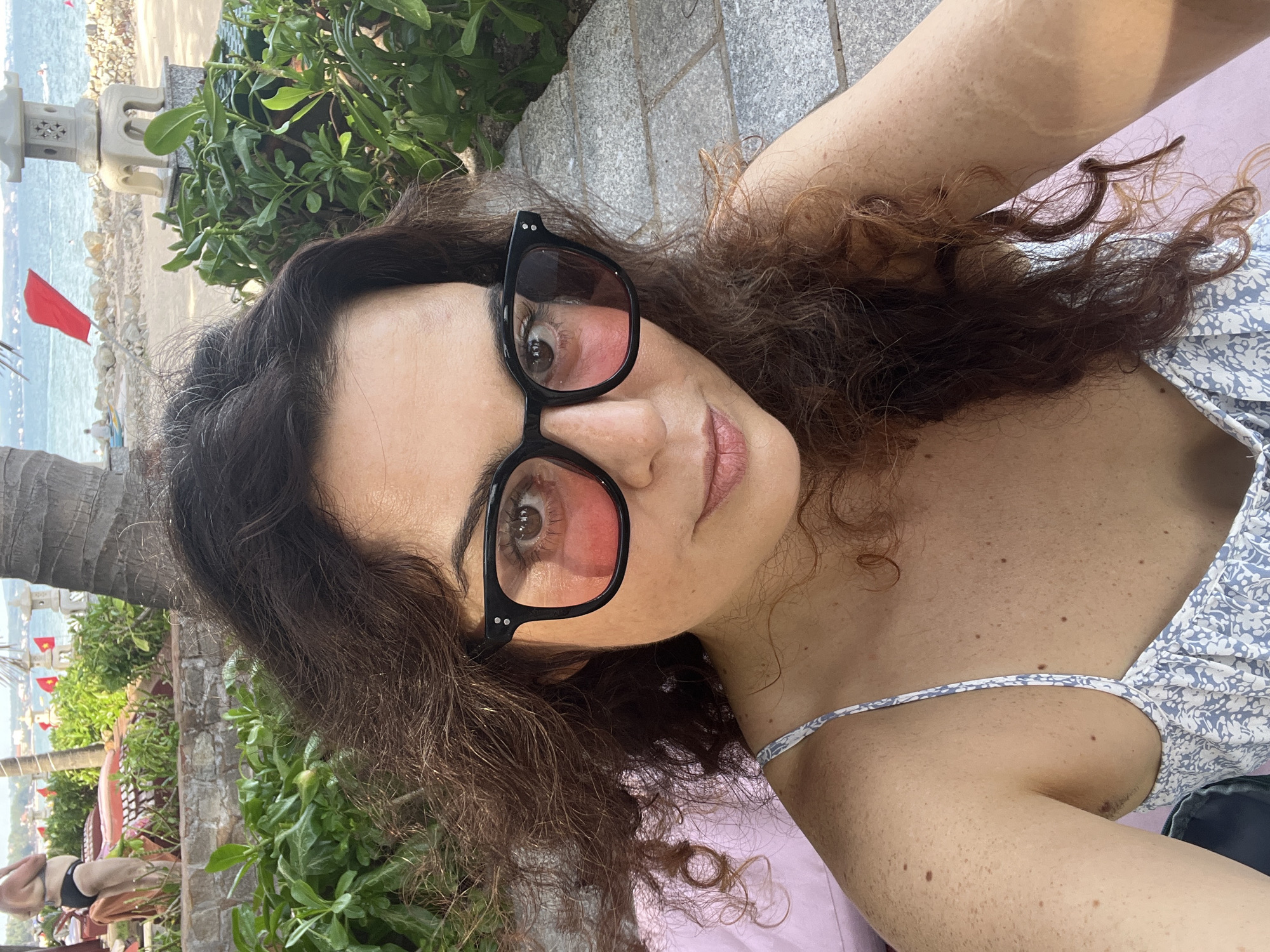 A woman with curly brown hair and black-rimmed glasses with pink lenses takes a selfie outdoors. She is wearing a light-colored dress with a floral pattern. Behind her, a stone pathway leads to a sandy beach with the ocean and a clear sky. Lush green foliage surrounds the pathway. In the distance, a person in a black bikini is visible near some lounge chairs and palm trees. Red flags are planted along the beach, and a stone lantern sits on a pedestal. The scene appears to be in Mũi Né, Vietnam, a coastal town known for its beaches and resorts.