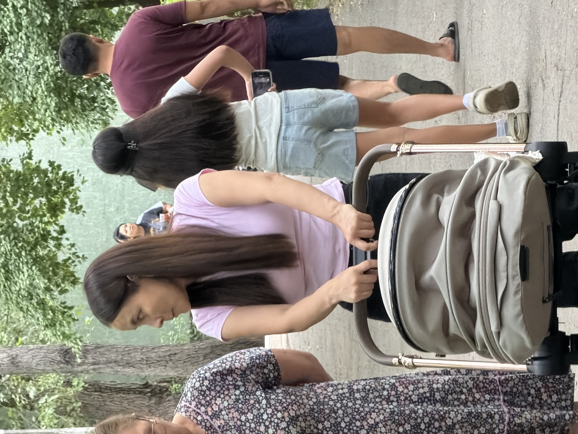 A group of people are outdoors, possibly in a park or along a pathway. In the foreground, a woman with long, dark hair, wearing a light pink t-shirt, pushes a light beige stroller. Her gaze is directed downwards. Behind her, a person wearing a floral patterned dress is partially visible. Further back, another woman with long dark hair, wearing a light blue t-shirt and denim shorts, is holding a smartphone. To her left, a person in a burgundy t-shirt and dark shorts is walking away from the camera. The ground appears to be paved or a similar hard surface, with some greenery and trees in the background. The lighting suggests it is daytime. No specific text is visible.