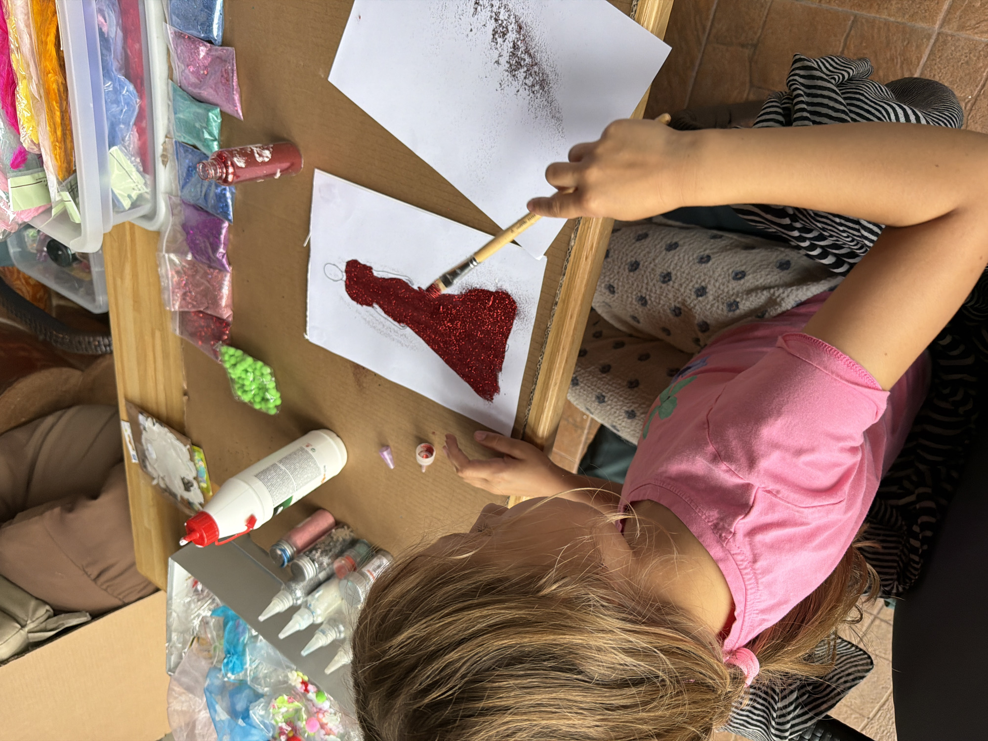 A young girl with blonde hair tied in a ponytail is sitting at a table and applying red glitter to a drawing of a dress with a paintbrush. She is wearing a pink t-shirt and patterned leggings. The table is covered with various craft supplies, including bottles of glue, packets of glitter in different colors, and containers of other craft materials. The scene appears to be outdoors, possibly on a patio or balcony, as indicated by the tile flooring visible in the lower portion of the image. The presence of craft supplies suggests a creative activity taking place. The image is taken from a slightly elevated angle, looking down at the girl and the table.