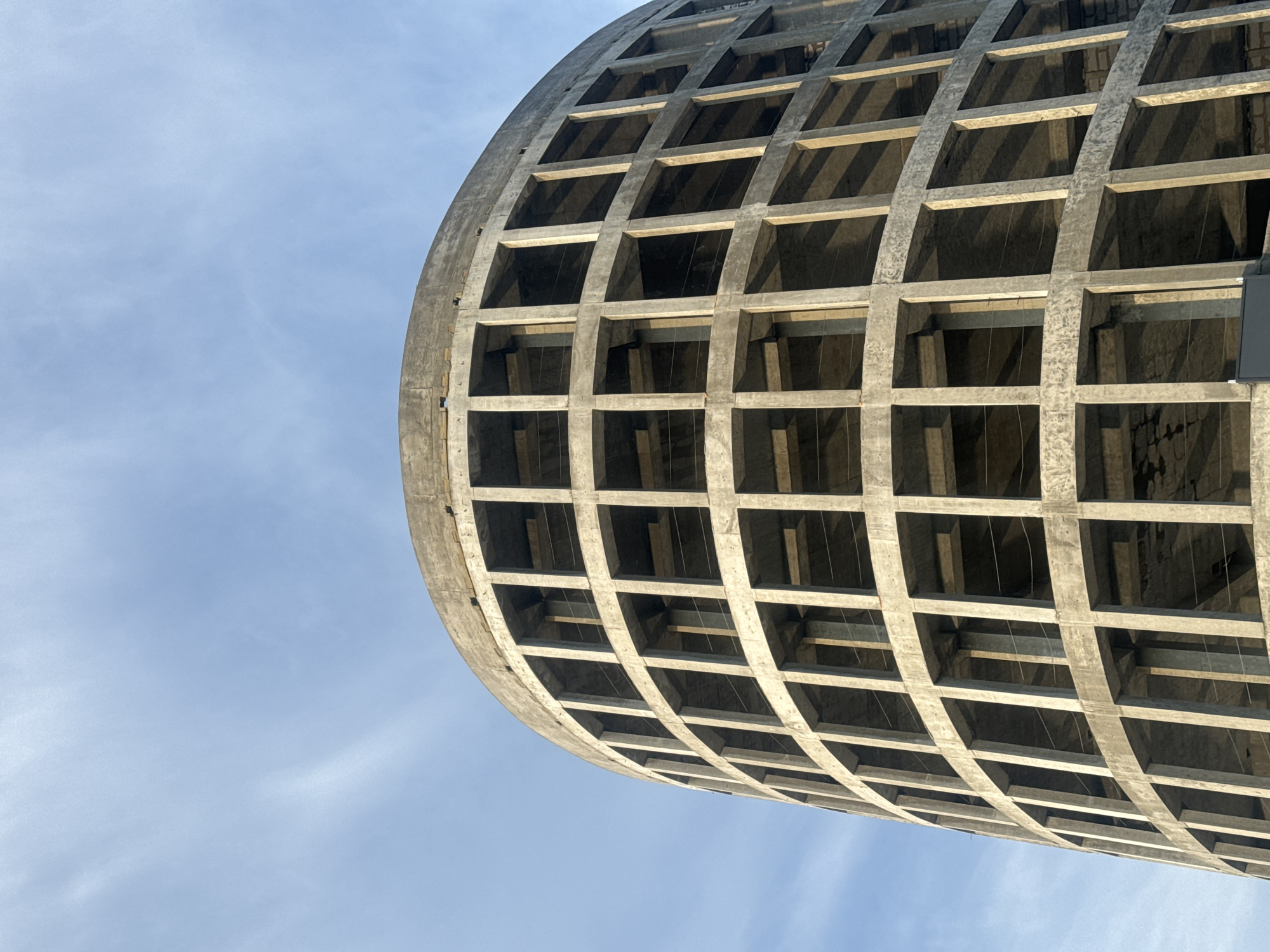 The image shows a partially constructed circular building with a concrete exoskeleton. The building appears to be a multi-story structure with numerous rectangular openings that will eventually form windows or balconies. The concrete is raw and unfinished, indicating an ongoing construction process. The sky above is a light blue with wispy white clouds, suggesting it's daytime, possibly late afternoon due to the soft lighting. The building's curvature is pronounced, and its internal structure, consisting of vertical supports and horizontal floor slabs, is visible through the openings. No people or other distinguishing features are present in the frame, focusing solely on the architectural form and the surrounding sky. The location is identified as Irkutsk, Russia, though no specific landmarks within the city are recognizable from this image.