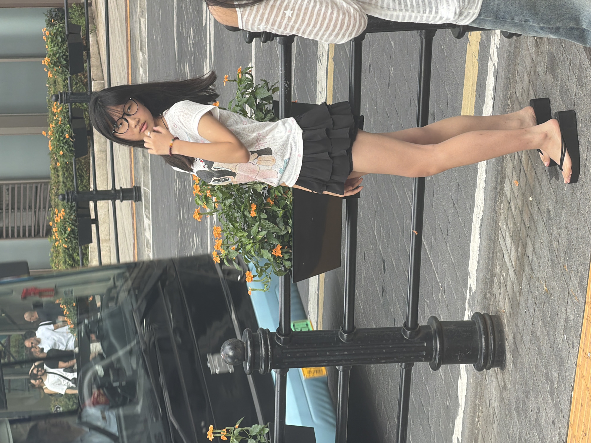 In Yuzhong District, China, a young East Asian female stands on a paved sidewalk. She is wearing a white t-shirt with a graphic print, a black ruffled skirt, and black platform flip-flops. She has long dark hair and is wearing glasses. She is looking towards the left side of the frame with her right hand touching her chin. Behind her, a black metal fence with decorative posts separates the sidewalk from a street with a blue bus. Orange flowers are planted in black rectangular planters along the fence. In the background, there are buildings and other pedestrians. The lighting suggests it is daytime.