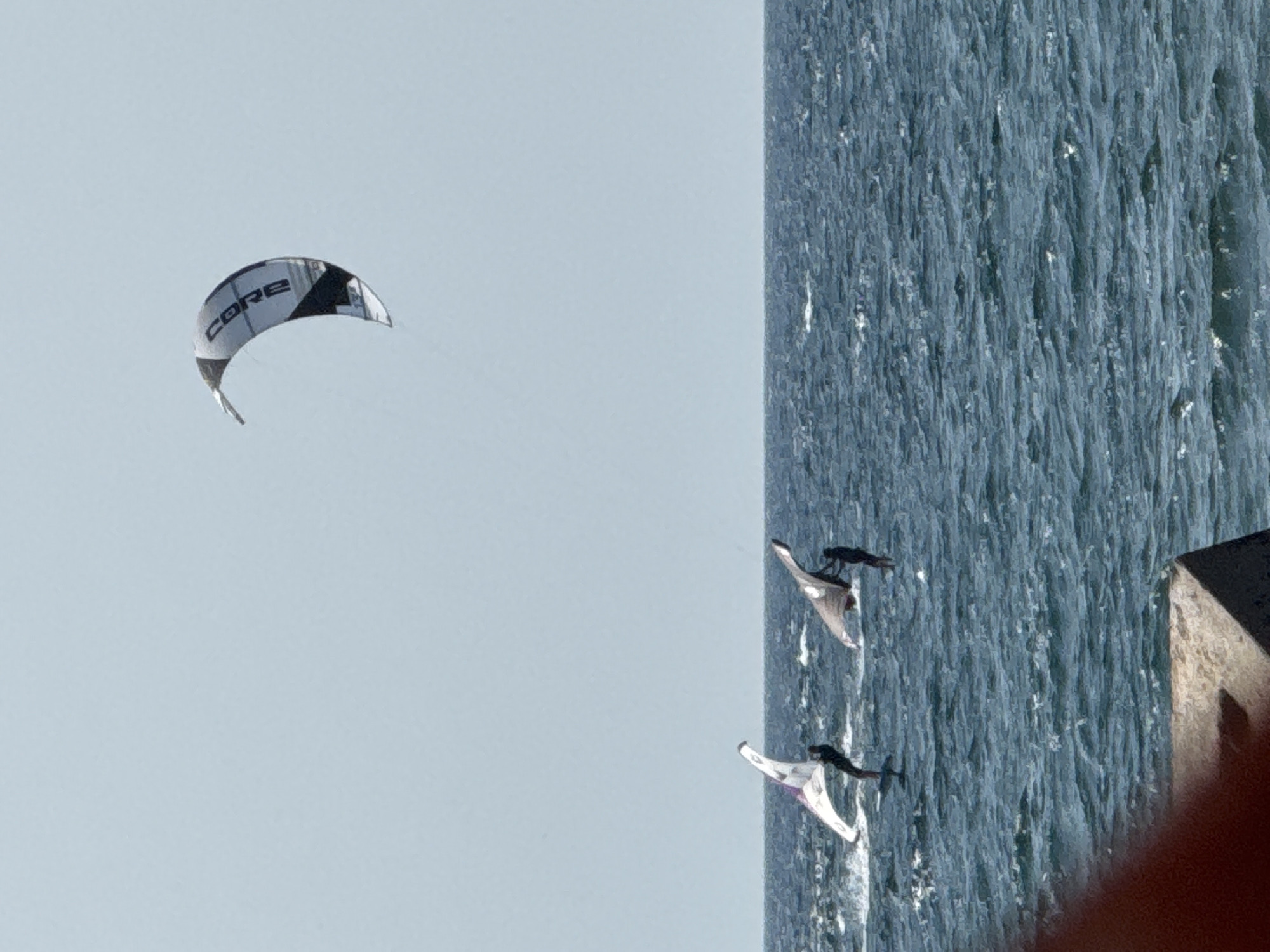 The image depicts a bright, sunny day over the sea, with a pale blue sky. In the foreground, a portion of a concrete structure is visible. The middle ground shows the choppy, dark blue ocean. Two individuals are engaged in watersports. On the left, one person is hydrofoiling, propelled by a white and purple wing. On the right, another person is also using a wing, this one grey, and appears to be standing on a board or hydrofoil. In the upper right quadrant of the image, a large, grey and white kite with "CORE" printed on it is soaring in the sky. The scene is likely coastal, given the presence of a structure and the open water. The location is specified as Sevastopol, Ukraine.