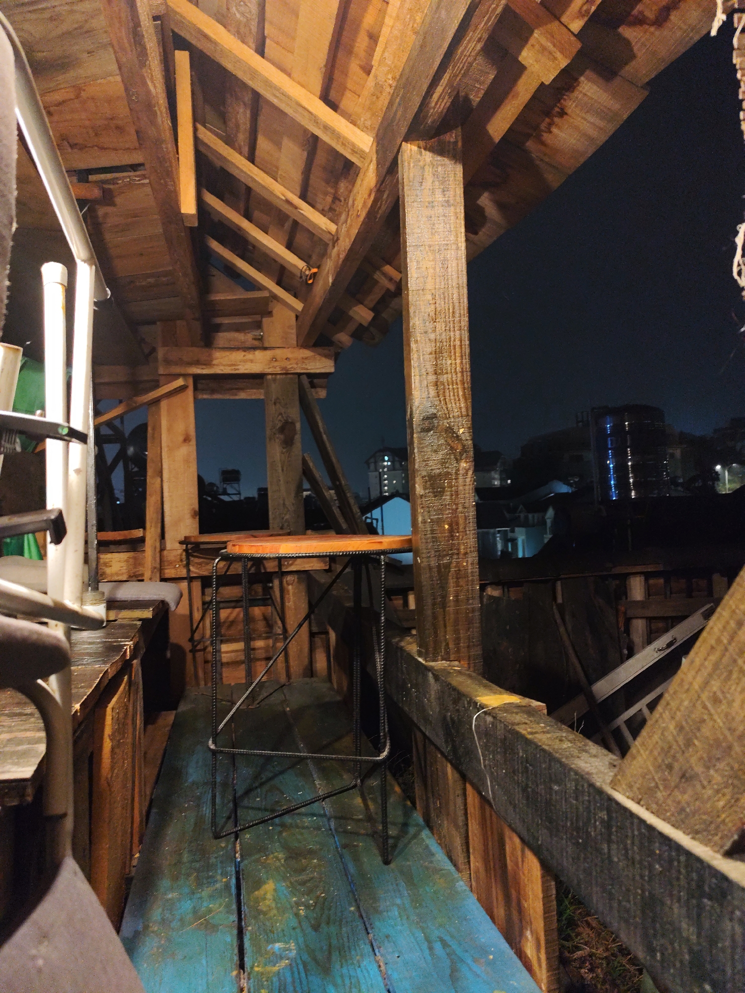 The image shows a partially enclosed wooden structure, likely a balcony or outdoor seating area, located in Da Lat, Vietnam. The structure is constructed from rough-hewn timber with a prominent roof overhead. A metal bar stool with a wooden seat sits on a blue-painted wooden floor. Outside the structure, the city lights of Da Lat are visible against the night sky. The lighting is dim, and the overall atmosphere is rustic. There is no evidence of any human interaction in the shot.