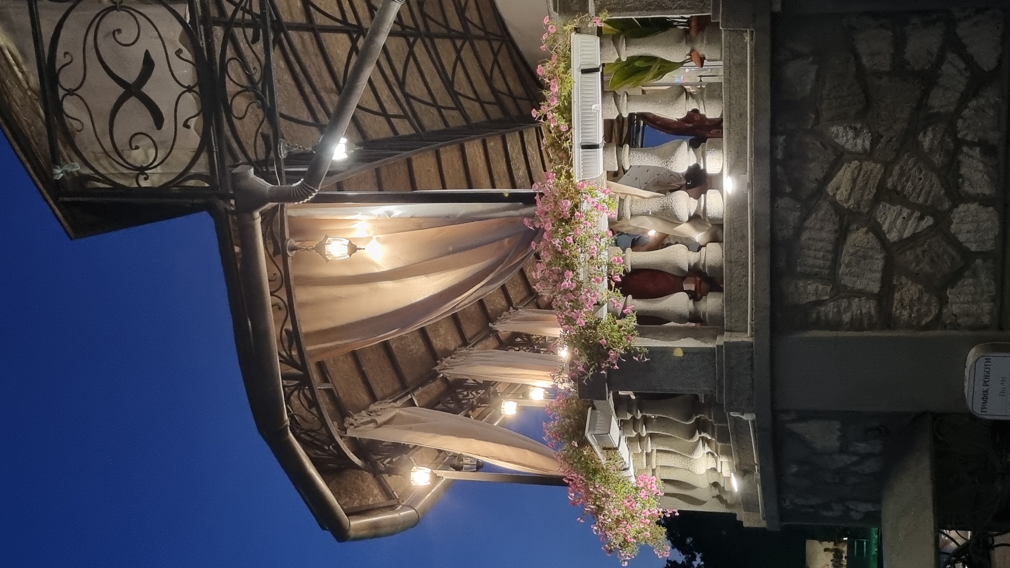The image shows a nighttime view of an outdoor balcony decorated with pink flowers in planters and ornate stone railings. The balcony is covered by a roof supported by a metal structure, with hanging lanterns providing illumination. The background is a deep blue twilight sky. The scene appears to be at an establishment in Berehove, Ukraine, possibly a restaurant or cafe, given the outdoor dining ambiance. The architecture suggests a blend of traditional and decorative elements.