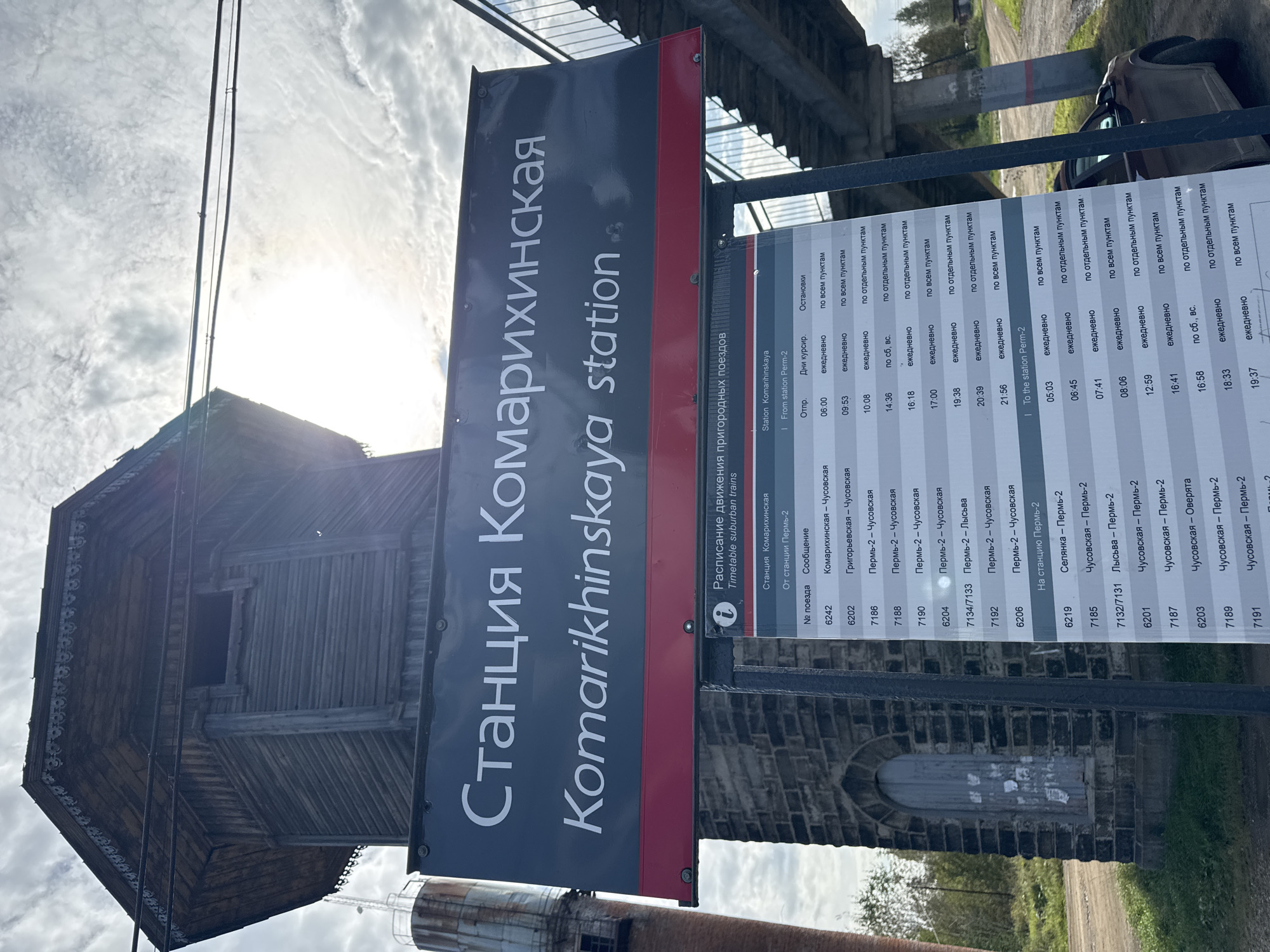 The image shows a weathered wooden water tower and a signpost in a rural setting, likely in Russia. The sign displays the name of a train station in both Russian and English: "Станция Комарихинская Komarikinskaya station". Below this, a timetable for suburban trains is visible, listing train numbers, destinations, departure times, and operating days. The surrounding environment appears to be overgrown with vegetation, and a car is parked in the background. The sky is cloudy.