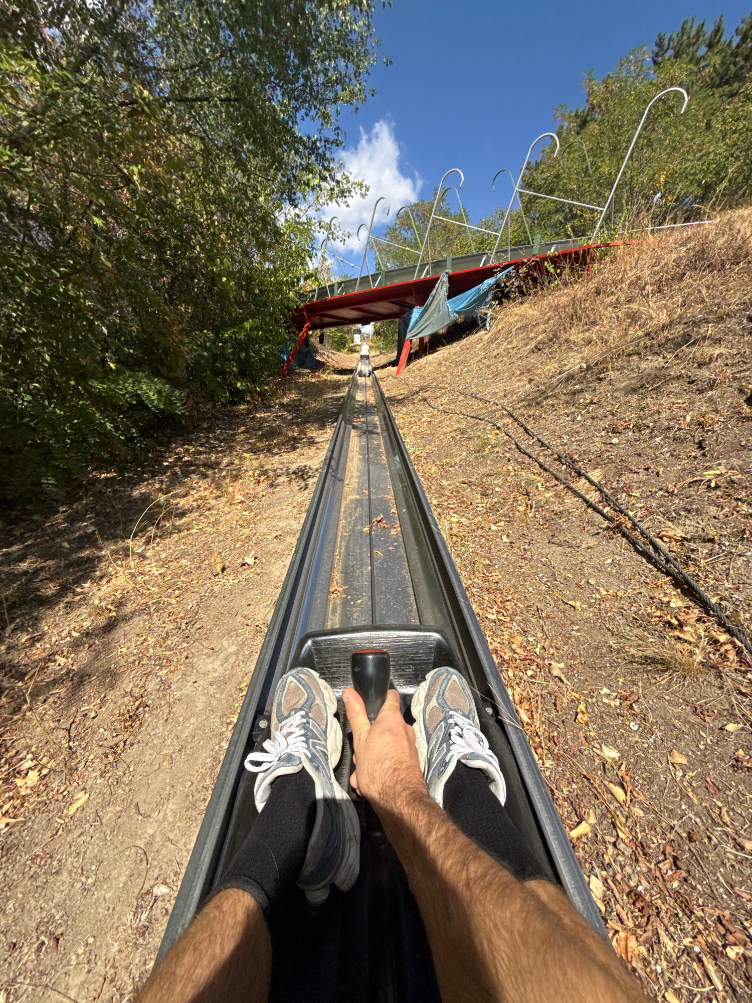 The image shows a first-person perspective of someone riding a toboggan down a track. The toboggan is a dark grey color and appears to be made of metal or plastic. The rider's legs and feet are visible, wearing white athletic shoes with grey accents and black socks. Their right hand is on a black handle in front of them, suggesting they are controlling the toboggan's speed or direction.
The toboggan track is a narrow, metal channel that descends a grassy, dry slope. The surrounding environment is characterized by dry grass, fallen leaves, and trees with green foliage. In the background, a red structure with white metal supports is visible, part of the toboggan run's infrastructure. The sky is blue with scattered white clouds.
This toboggan run is located at the Budapest Children's Railway, also known as the Pioneer Railway (Gyermekvasút), which is situated in the Buda Hills of Budapest, Hungary. The railway is operated by children and is a popular tourist attraction. The image captures a sunny day with ample natural light, highlighting the textures of the natural environment and the man-made toboggan structure.