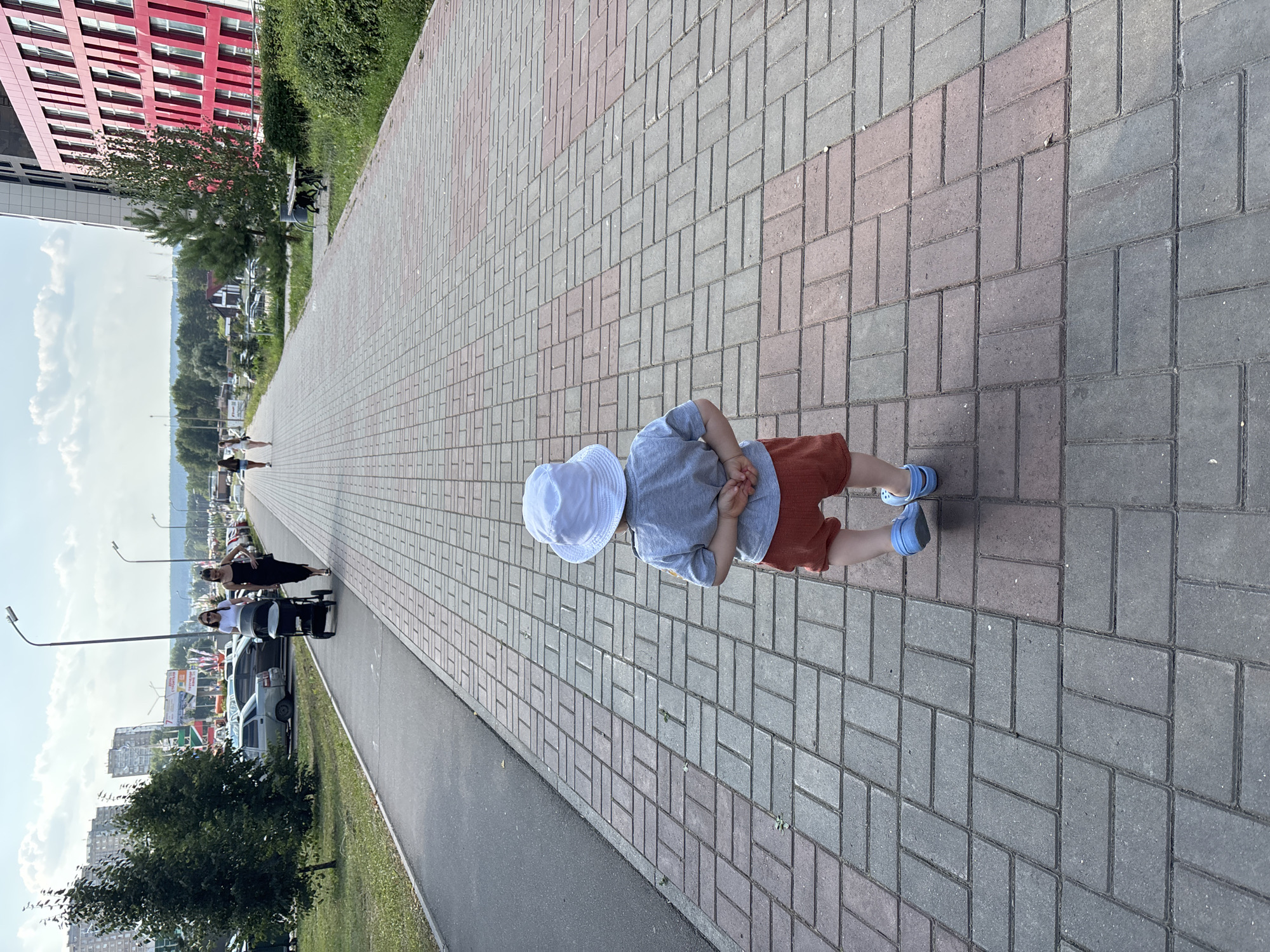 A young child wearing a white bucket hat, grey t-shirt, and orange shorts walks away from the camera on a brick-paved sidewalk. The child wears blue Crocs. The sidewalk is lined with grass and trees on the left, and a modern red building with many windows on the right. In the background, several other people are walking on the sidewalk, including a woman pushing a stroller and another woman walking behind her. Cars are parked along the left side of the sidewalk. In the distance, a cityscape with buildings and a treeline is visible under a cloudy sky. This scene is located on the embankment of Naberezhnye Chelny, Russia, specifically near the Chelny-Gorod park.
