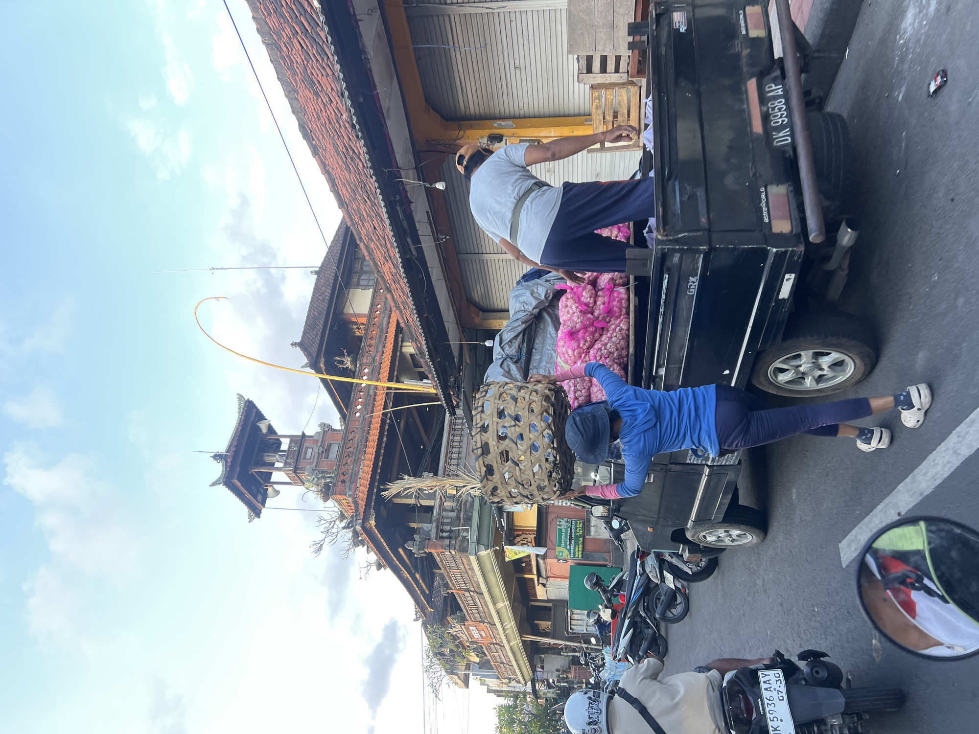 In Denpasar, Indonesia, on a street lined with traditional Balinese architecture, a black flatbed truck is parked. Two individuals are interacting with the truck's contents. One person, wearing a blue hooded jacket and a dark hat, is carrying a large woven basket on their head and is positioned behind the truck. The other person, wearing a grey t-shirt and dark pants, is bent over inside the truck bed, seemingly engaged with items loaded onto it. The truck bed contains large sacks filled with pink-colored produce, possibly onions or garlic, and a tarp covering other items. A person on a motorcycle, viewed from behind, is in the foreground on the left side of the image, with their motorcycle license plate visible. The street is paved, and parked motorcycles are visible on the left side of the road. The architecture in the background features ornate rooftops and balconies, typical of Balinese buildings. The sky is partly cloudy.