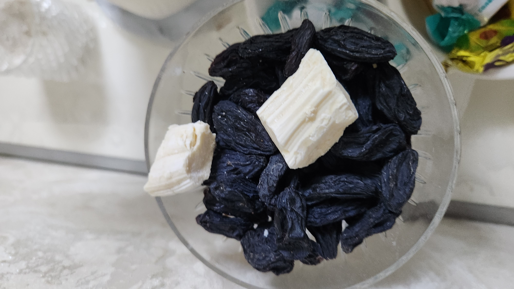 A close-up, overhead shot shows a clear glass bowl filled with dark raisins. Two rectangular pieces of white candy or chocolate are placed on top of the raisins, one near the top center and the other slightly lower and to the right. The background is out of focus, with a textured, light-colored wall and hints of other objects. The scene is indoors.