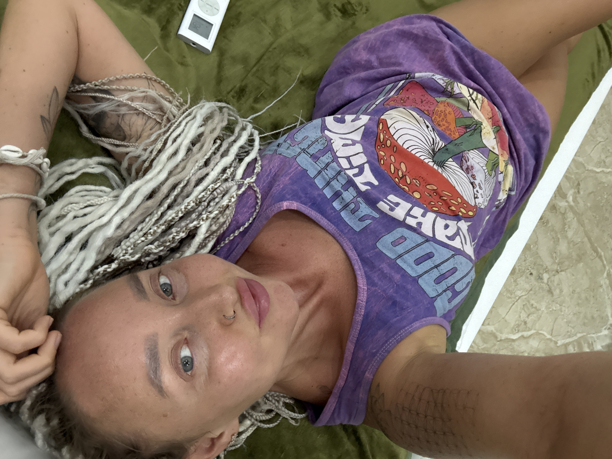 A person with blonde braids is lying on a green surface. They are wearing a purple tank top with a graphic of mushrooms on it and text that appears to say "GOOD VIBES". A white iPod is visible in the upper left corner of the image. The person has several tattoos on their arms. The lighting suggests it is daytime, and the scene appears to be indoors, possibly a bedroom or living area.