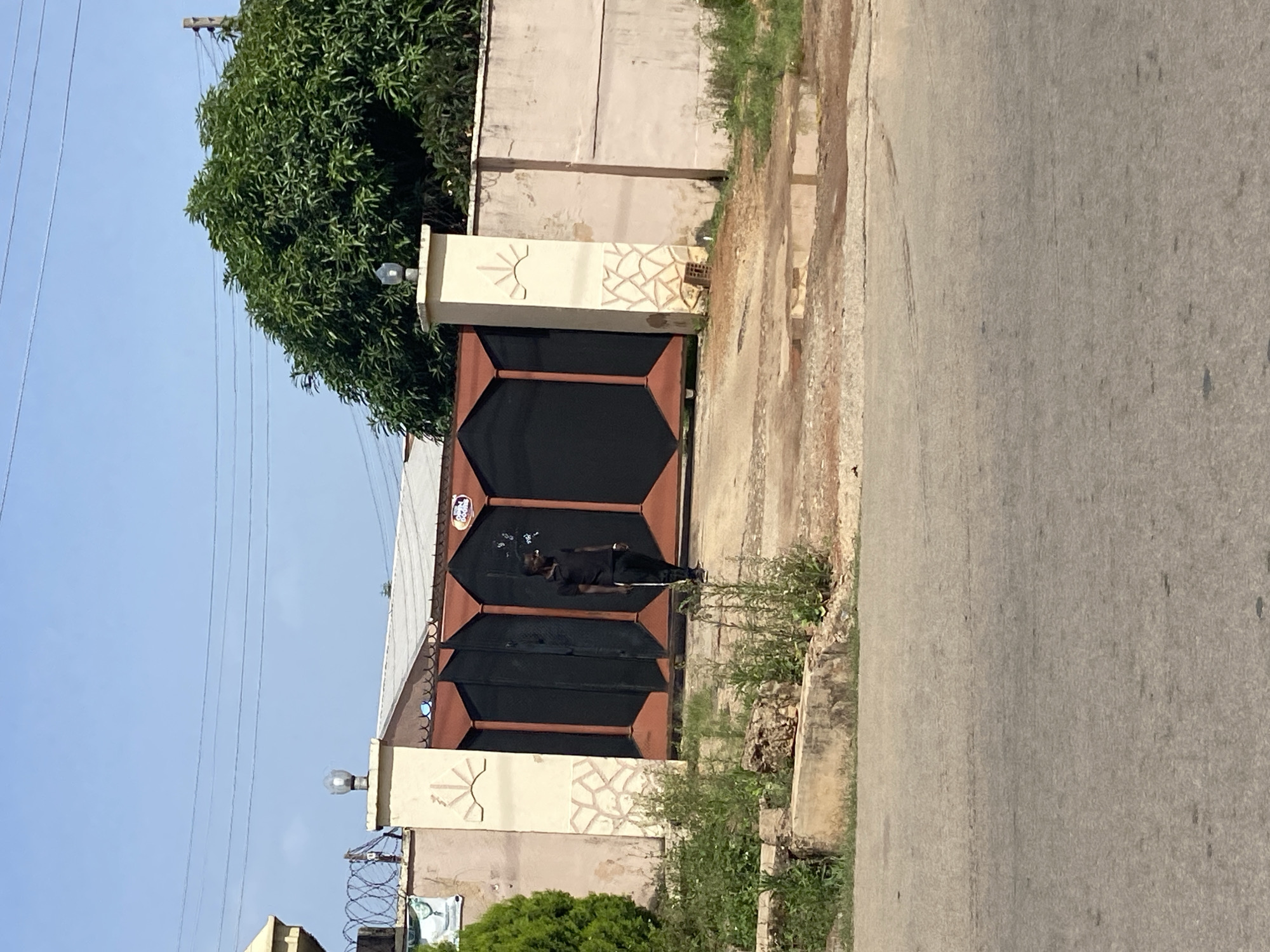 The image depicts a street-level view of a closed, dark-colored metal gate with reddish-brown trim, located in Jos, Nigeria. A man dressed in a black short-sleeved collared shirt and dark pants stands to the left of the gate, holding a stick or pole. The gate is flanked by two pillars, the left one adorned with a stylized sunburst emblem and a geometric pattern, and the right one with a similar sunburst emblem and a more abstract pattern. Behind the gate, a concrete wall extends to the right, partially obscured by a large, lush green tree. Barbed wire runs along the top of the wall to the left of the gate. The ground in front of the gate is unpaved and shows signs of wear, with some patchy vegetation and a small puddle of water. A paved road with tire marks is visible in the foreground. The sky is clear blue.