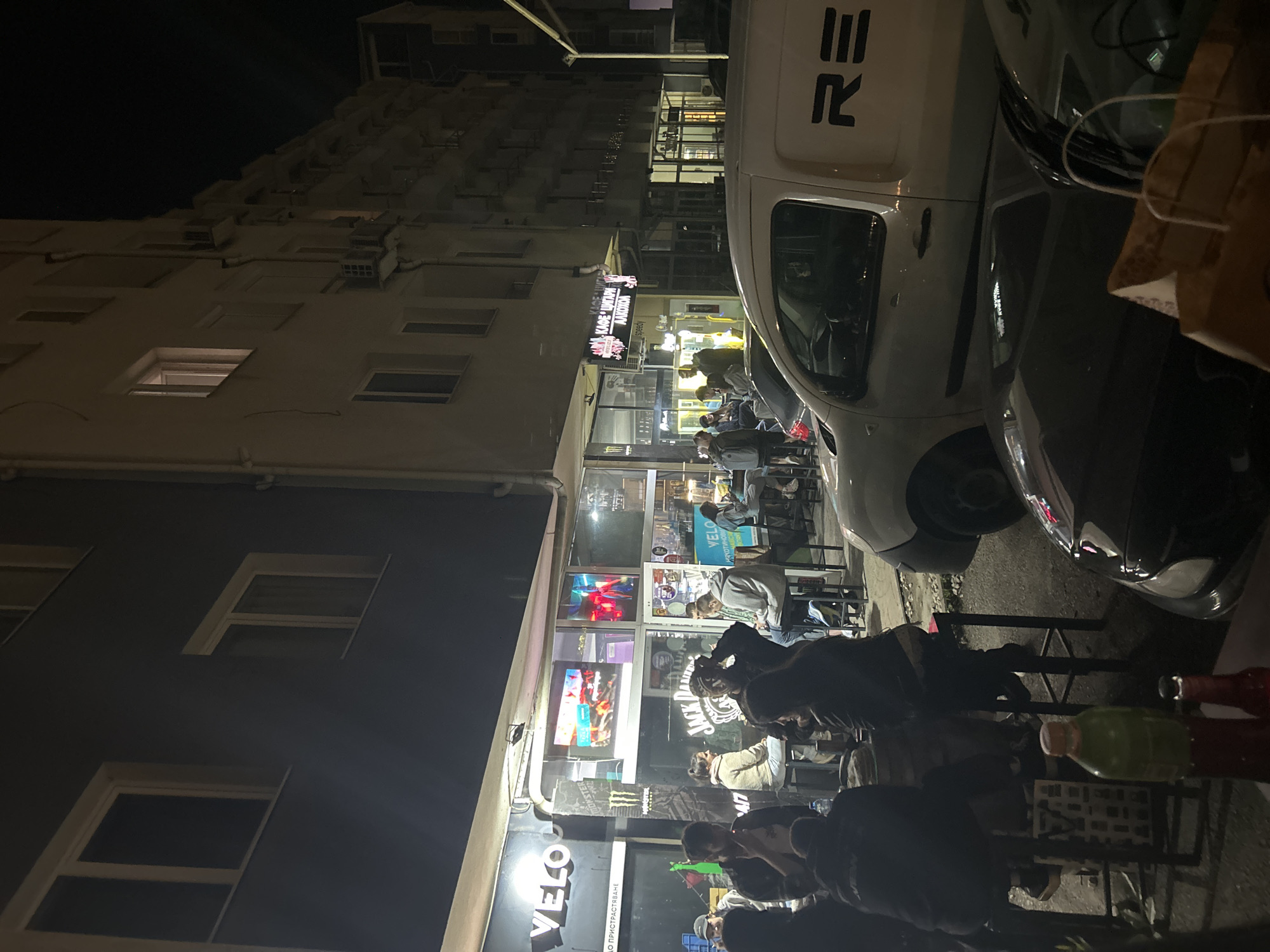 This media file, set at night in Sofia, Bulgaria, captures a bustling street scene outside a commercial establishment. Multi-story buildings line the background, with windows and air conditioning units visible on their facades.
The primary subjects are a group of people gathered outdoors, seated at small, dark tables with chairs. They appear to be socializing in a casual setting. Visible objects on the tables include bottles, one green and one red, suggesting beverages.
Two vehicles are prominently parked on the street: a white van and a darker car next to it. The van bears a partial black logo or text that reads "≡R". A brown paper bag rests on the dashboard of the darker car.
The establishment features several signs and logos. A main, brightly lit sign displays "KAΦE ШΟΠ AKOTIKA" (Coffee Shop Akotika) with "speedy" written below it. Further down, on the storefront glass, a white "VELO" logo is visible with the Cyrillic text "СВОБОДНО ПРИСТРАСТЯВАНЕ" (Free Addiction/Freely Addictive) underneath. Inside the establishment, signs for "Jack Daniel's" and "Monster Energy" are discernible. Two televisions are also visible through the window, displaying colorful graphics.
The time of day is clearly night, with artificial lights illuminating the storefront and the gathering. The overall mood appears relaxed and social, characteristic of an evening hangout spot. No specific weather conditions are evident.