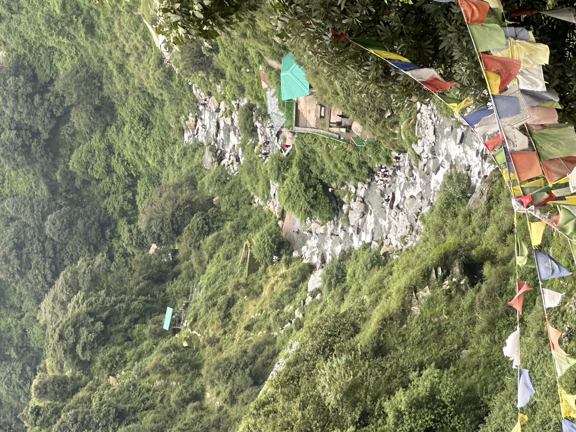 This image depicts the Bhagsu Waterfall in Bhagsu, India, a popular tourist destination nestled within a lush, green mountainside. A river, characterized by its rocky bed and flowing water, carves its way through the steep terrain. Numerous individuals are visible along the riverbanks and in the water, suggesting recreational activity. Several structures, including small huts with green roofs and a more complex building with a red and green roof, are present, indicating developed areas for visitors. Prayer flags, a common sight in the region, are strung across the foreground, adding a cultural element to the natural landscape. The overall environment is one of dense vegetation, with trees and bushes covering the slopes of the valley.