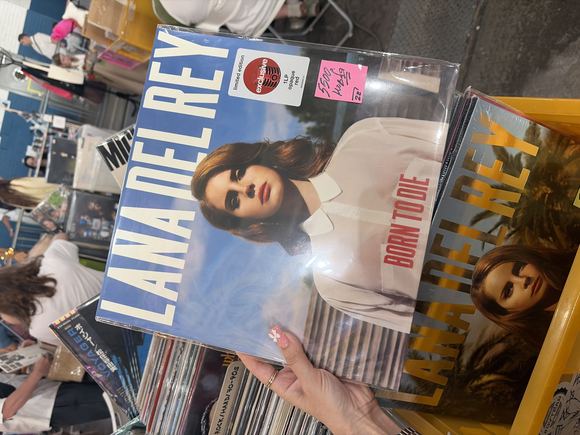 An individual's left hand holds up a vinyl record of Lana Del Rey's "Born to Die" album at a record fair in Saint Petersburg, Russia. The album cover features Lana Del Rey centrally, with "LANA DEL REY" in white text at the top and "BORN TO DIE" in red text on her white collared shirt. The record is encased in clear plastic, displaying a white "limited edition exclusive Target" sticker indicating a "1LP opaque red" pressing. A pink sticky note attached to the plastic reads "5500" and "Новая" (Russian for "New") in black marker, followed by "28'", likely indicating a price in rubles and vendor identification.
The hand is light-skinned, with manicured pink fingernails adorned with white polka dots and a small white flower appliqué on the ring finger. Two thin gold rings are worn on the ring finger. Below the held record, in a yellow plastic bin, another copy of "Born to Die" with gold lettering is partially visible. To the left, a stack of vinyl records shows spine labels, including one indicating "ROCK/HARD/70-90s". Another record further left displays "VOYAGER" with Japanese text.
The background reveals a busy indoor setting, consistent with a record market or convention. Multiple blurred individuals, casually dressed, are visible, some appearing to browse at stalls. Metal barriers, display tables with merchandise, and overhead fluorescent lighting are discernible. The flooring is a light-colored, possibly concrete, industrial surface.