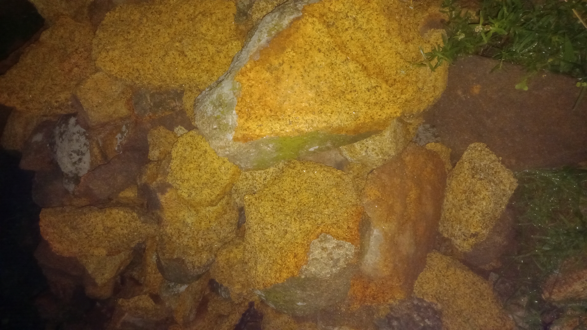 A close-up, eye-level, medium shot captures a pile of large, rough-textured rocks in Katon Rikkos, Nigeria. The rocks vary in color, with many exhibiting a prominent yellow-orange hue, some showing patches of light gray and dark brown. Some rocks are speckled with lighter colored particles, giving them a granular appearance. A few areas on the rocks show a greenish moss or lichen. The ground beneath and around the rocks is dark and appears wet, with some sparse green grass visible in the lower right and bottom left portions of the frame. The lighting is dim, suggesting it may be nighttime or a heavily shaded area, casting shadows and creating a somewhat muted color palette. There are no people or animals visible in the image. The scene is devoid of any identifiable human-made structures or signs of recent activity. The composition is dominated by the textures and colors of the rocks, creating an abstract and somewhat rugged aesthetic.