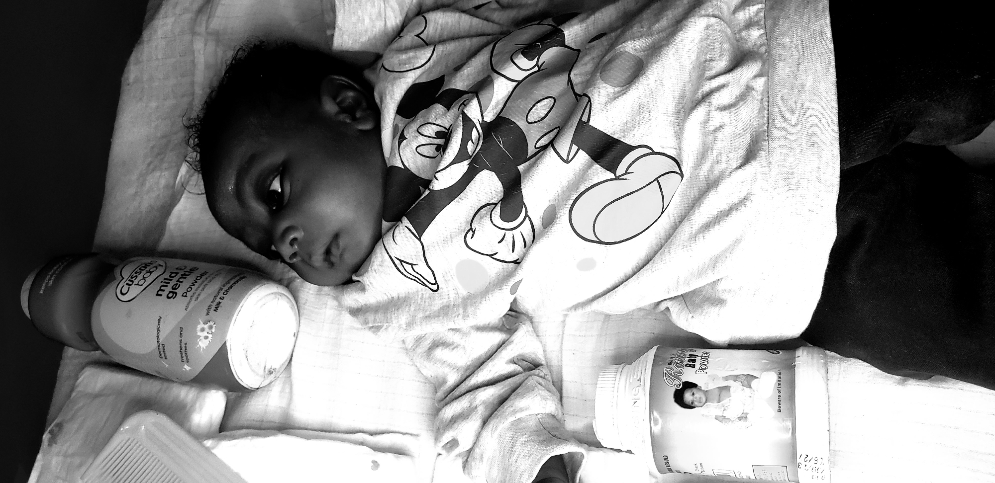 A baby lies on their back in a bed or cot. They are wearing a Mickey Mouse patterned shirt and dark trousers. Two bottles of baby powder are on either side of them. One is "Cussons Baby mild & gentle Powder" and the other is "Ratin Baly Powder." The location is Narayi, Nigeria.