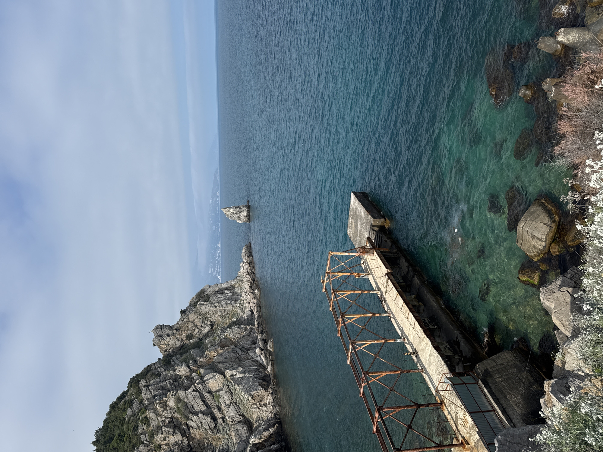 The image shows a view overlooking the Black Sea in Ливадийский поселковый совет, Ukraine. A rocky coastline is visible in the lower left, featuring layered grey rock formations and sparse vegetation. To the right of the rocks, a concrete pier or structure extends into the water. The structure has a rusty metal framework above and adjacent to it. The water near the coastline is clear with visible underwater rocks and vegetation. Further out, the sea is a deep blue color. In the distance, another rock formation rises from the sea. The sky is light blue with faint clouds.