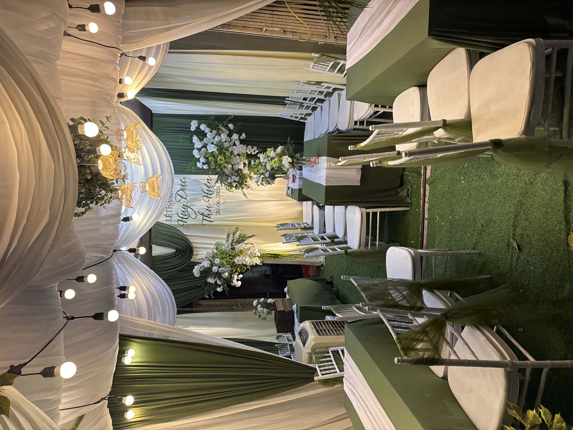 The image depicts a wedding reception setup in Hà Nội, Vietnam. The scene is decorated with white and green drapes, strung lights, and floral arrangements. Several tables draped in green are set up with white runners, and white chairs with green sashes are arranged around them. A cooler is visible near one of the tables. A large floral arrangement is placed near a sign with Vietnamese text and a date, "20.06.2026". The overall ambiance suggests an outdoor or semi-outdoor event.