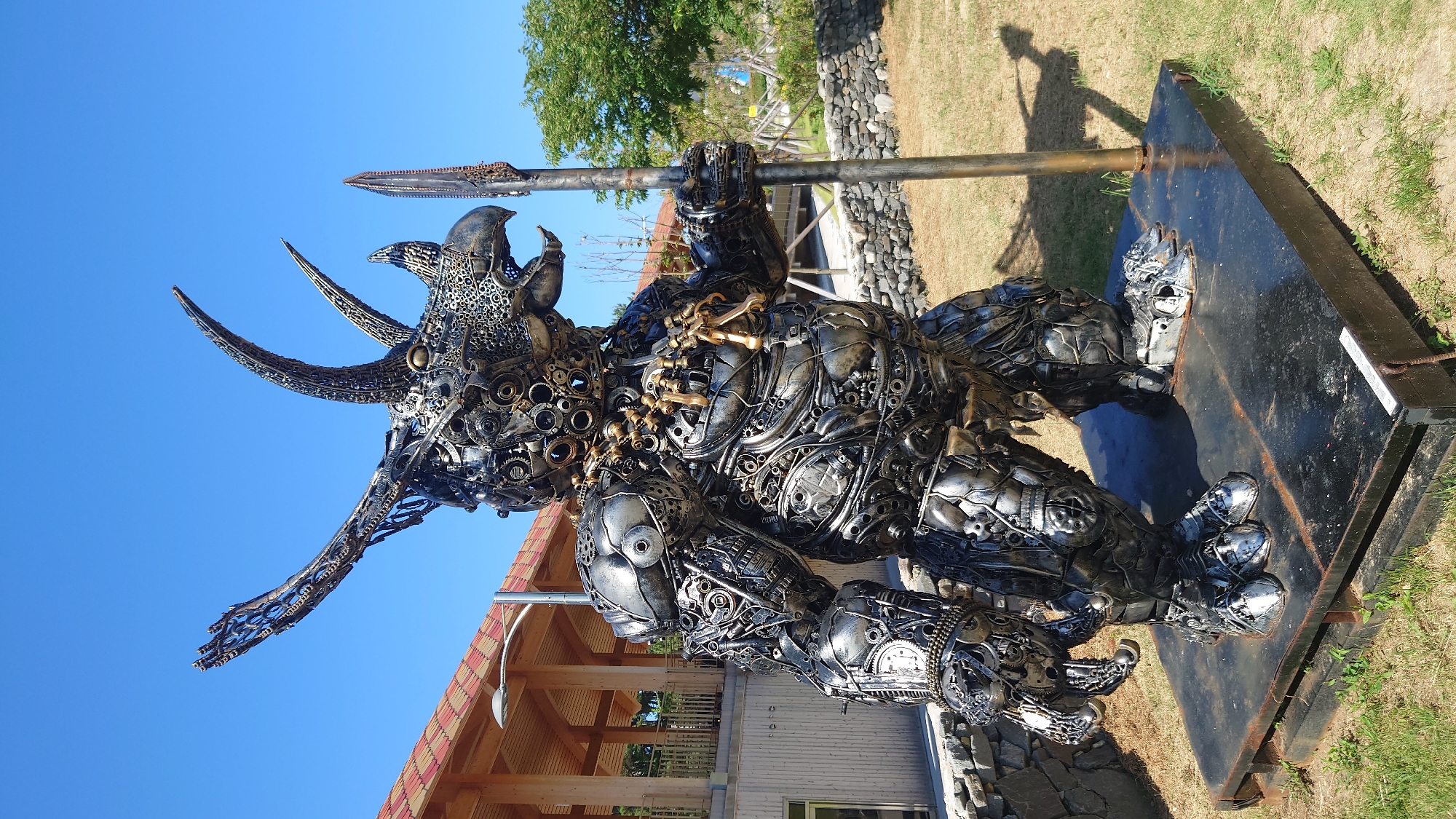 An outdoor, full-shot photograph shows a large, metallic sculpture of a dinosaur, likely a Triceratops, with a bird-like head. The sculpture is made from various recycled metal parts, including gears, chains, and car parts, giving it a steampunk aesthetic. It stands on a black metal platform in a grassy outdoor area. The dinosaur sculpture is holding a long, metal spear in its right hand. In the background, there is a wooden building with a red tiled roof and a clear blue sky. The location is the Ulsan Grand Park in Ulsan, South Korea, specifically within the park's sculpture garden. The lighting suggests it is daytime, with a bright sun casting shadows.