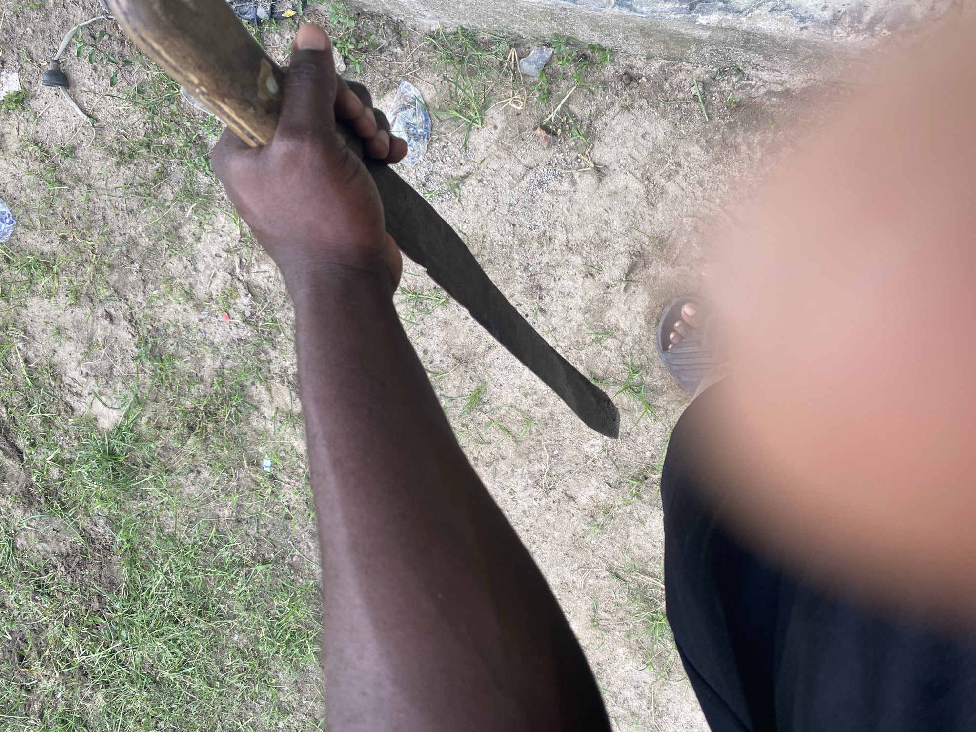 A person with dark skin is holding a dark-colored machete. The person is wearing black pants and a sandal on one foot. The background consists of dirt, grass, and a concrete surface. The image is taken from a high-angle perspective.