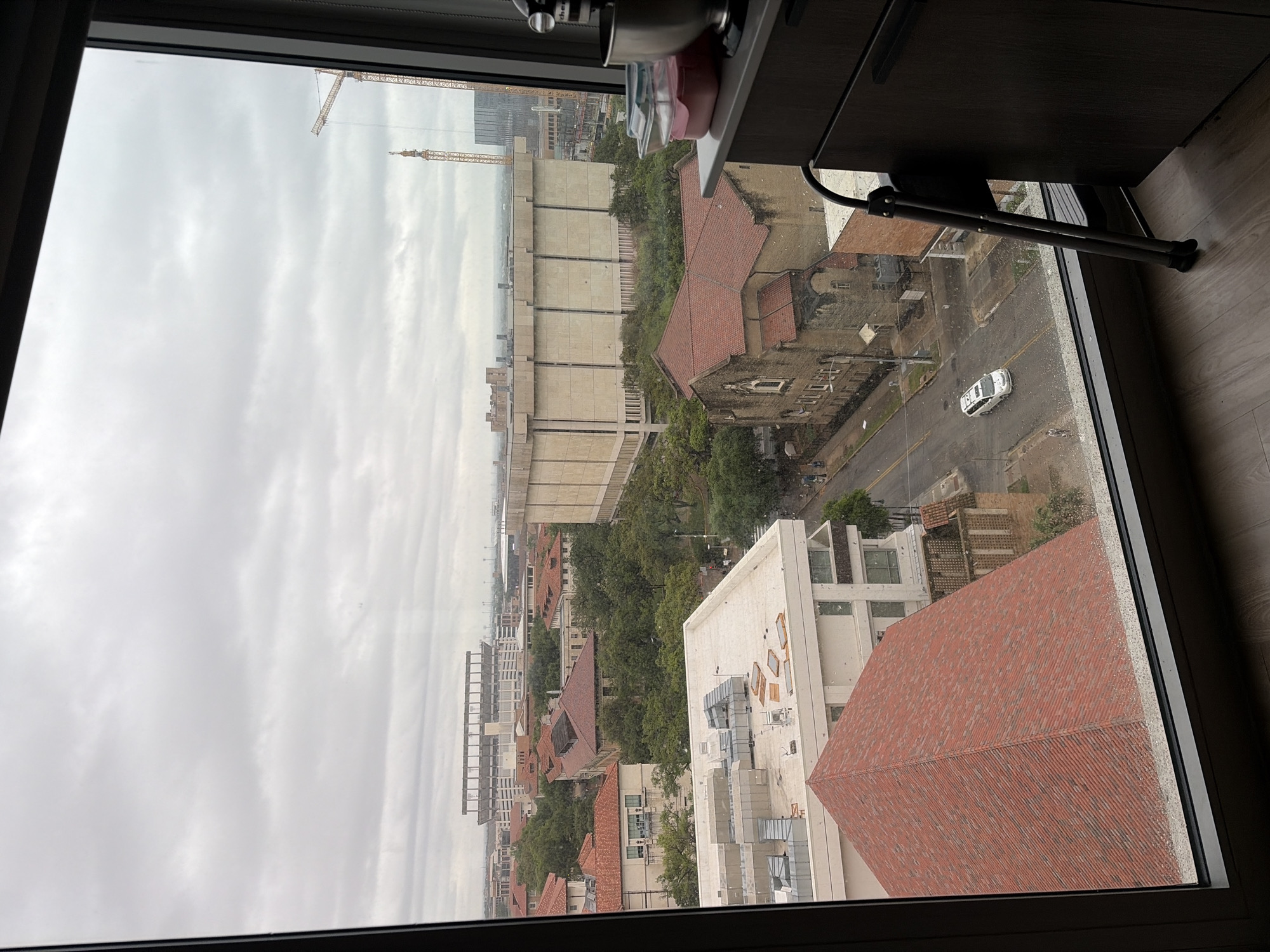 View from a high-rise building in Austin, Texas, overlooking the University of Texas at Austin campus. The focus is on the architectural landscape: UT Tower can be seen in the middle distance. Buildings, including red-tiled rooftops and modern structures, are visible. Cranes are in the distance, indicating construction. A road with a white vehicle is in the immediate vicinity. A gray, overcast sky fills the view. The image is framed by a window.