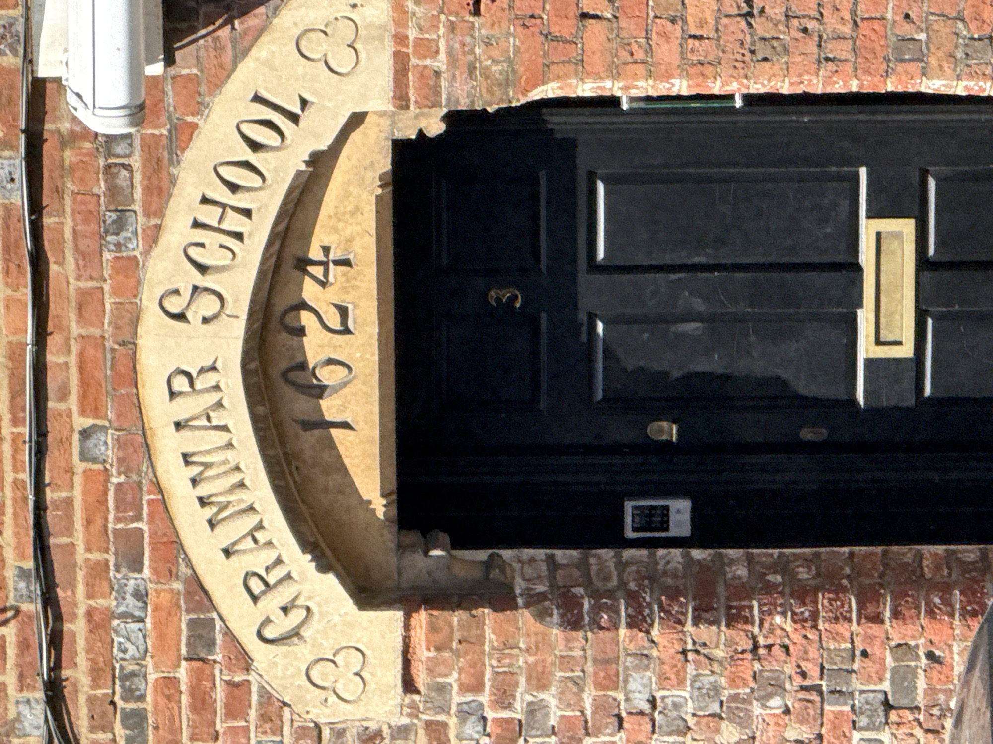 The image shows the entrance to Amersham Grammar School, located in Amersham, United Kingdom. The entrance is marked by a stone arch above the door, inscribed with "GRAMMAR SCHOOL" and "1624." The door is black and features a brass-colored mail slot and other hardware. The surrounding wall is made of brick. The photograph is taken at an angle, and the overall lighting suggests it's daytime.