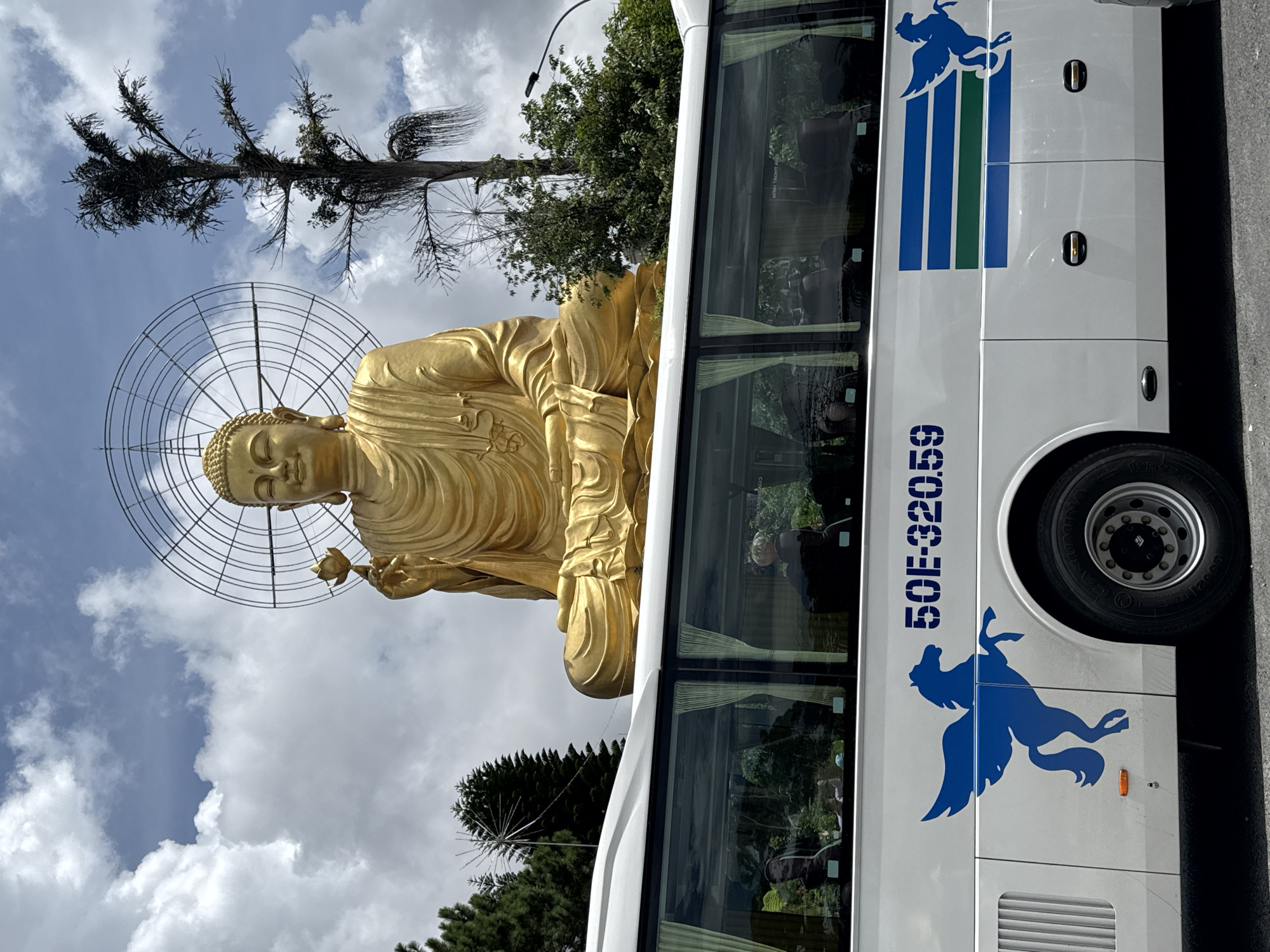 A monumental golden Buddha statue sits atop a hill, with a circular halo-like structure behind its head. In the foreground, a white bus with blue and green stripes runs across its side. The bus has a blue silhouette of a winged horse. The license plate on the bus reads "50E-32059". The bus's windows show reflections of trees and the sky, and some passengers are visible inside. The Buddha is holding a lotus flower. The environment appears to be a tourist attraction, likely in a mountainous area given the trees and clouds. The scene suggests a travel or pilgrimage context, with the bus serving as transportation to the site of the Buddha statue. The location is identified as Lâm Viên - Đà Lạt, Vietnam.