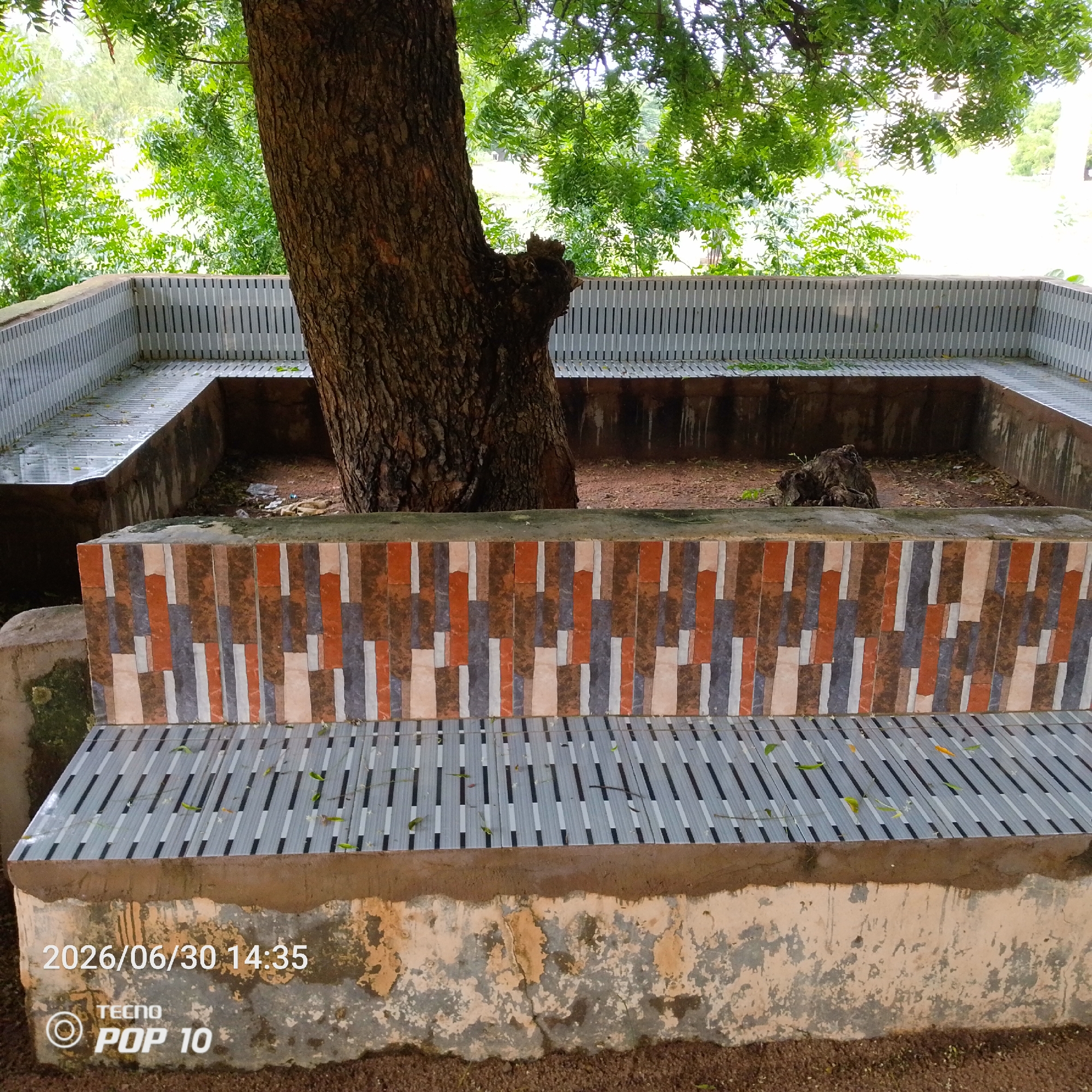 This is a horizontal, medium-shot, eye-level photograph taken in Jalingo, Nigeria. The scene depicts a tree with a rough, brown trunk in the center, surrounded by a built-in seating area. The seating is made of concrete and features a patterned tile facade in shades of brown, orange, and grey. The seating area itself is constructed with slatted metal benches. Beyond the seating, the background shows lush green foliage and a hint of a bright, overcast sky, suggesting daytime. There is no specific activity or event captured, the image appears to be a still life of the seating area under the tree.
Visible text in the lower left corner includes a timestamp "2026/06/30 14:35" and "TECNO POP 10" with a circular logo. The overall impression is one of a public or outdoor space that is somewhat unkempt, with fallen leaves scattered on the benches and debris visible in the central pit of the seating area. The concrete base of the seating shows signs of wear and tear, with peeling paint and staining. The presence of the TECNO POP 10 branding suggests the photo was taken with a mobile phone.