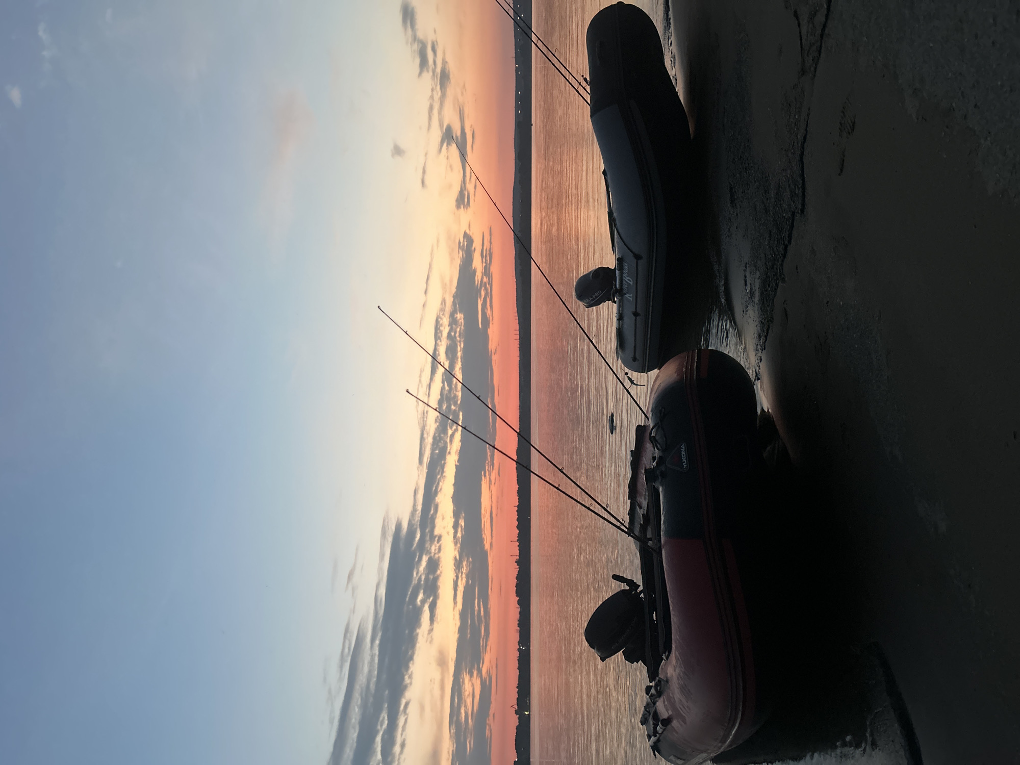 Two inflatable fishing boats are beached on the shore of the Kama River at sunset. The boat in the foreground is red and black and has a black outboard motor. The boat in the background is dark gray and has a black outboard motor. Both boats have fishing rods protruding from them. The sky is a gradient of blue, orange, and pink, with scattered clouds. The water is calm and reflects the colors of the sky. The city of Chaikovsky, Russia, is visible in the distance along the horizon.