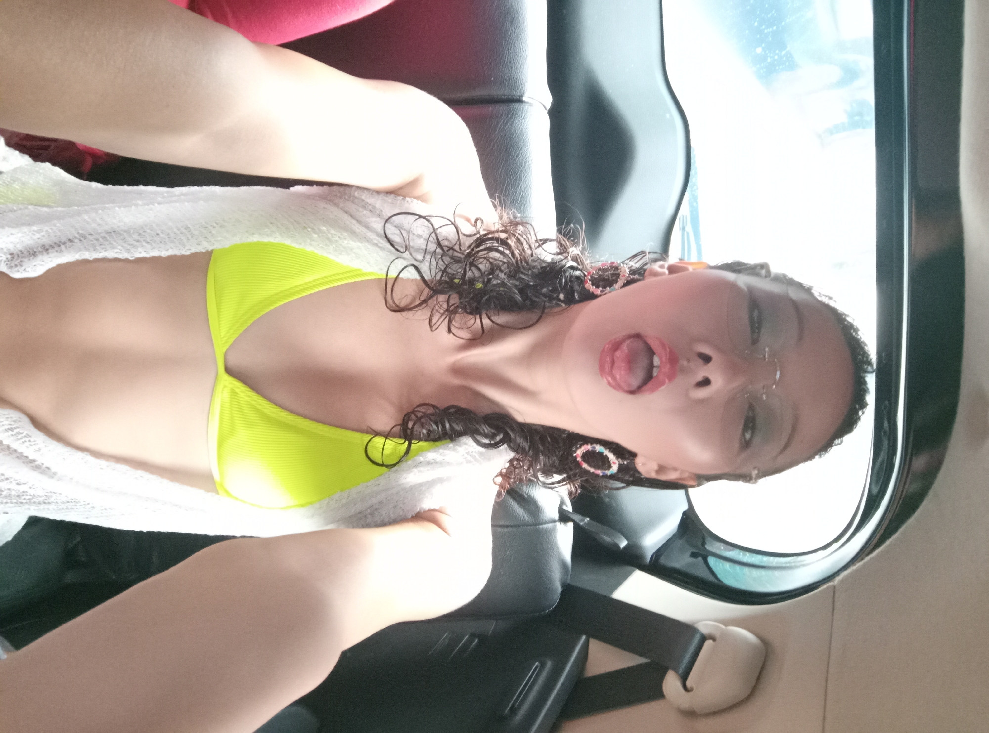 A woman is positioned in a car seat, wearing a neon yellow bikini top and a white knit cardigan. She has curly, dark hair adorned with multicolored hair accessories. Her mouth is open, tongue visible. The interior of the car is visible, with a black leather seat and a seatbelt buckle in view. Through the car window, a bright, undefined background can be seen. The image is angled, giving a distorted perspective. The location, based on the query, is North Kuta, Indonesia, though no specific elements in the image definitively confirm this.