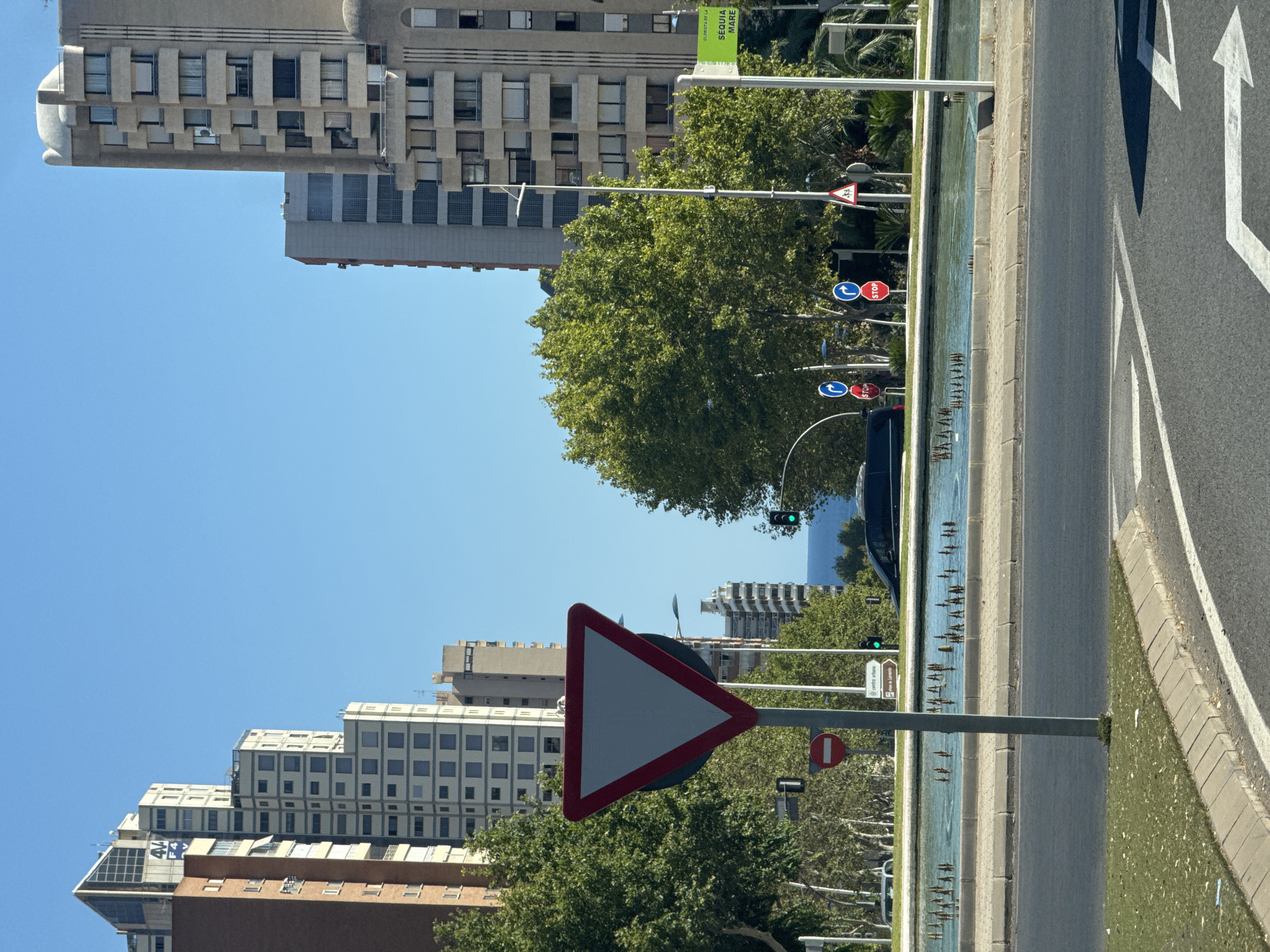 The image depicts a street scene in Benidorm, Spain, likely near the coast given the glimpse of the Mediterranean Sea in the distance. Tall, modern apartment buildings with many windows dominate the skyline, interspersed with lush green trees and a public fountain with water jets. Traffic signals are visible, including a green light, and several road signs, including a prominent give way sign, a "no entry" sign, and two "stop" signs. A black minivan is parked in front of the fountain. The foreground shows a street with white lane markings and a grassy verge. The sky is a clear, bright blue.