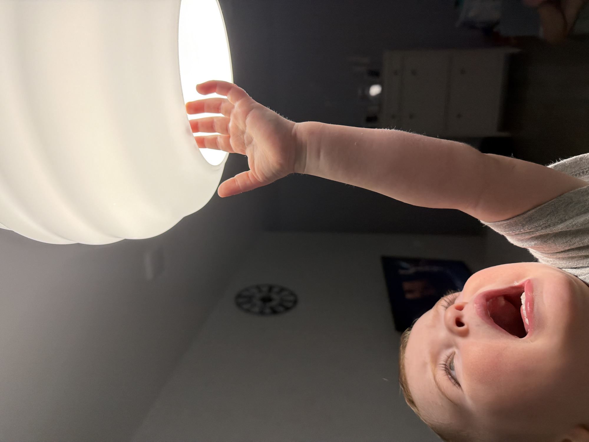 A baby, viewed from below and rotated upside down, reaches upwards towards a bright, frosted glass light fixture. The baby's mouth is open, suggesting a vocalization. The baby is wearing a grey garment. In the background, a wall with a clock and a picture is visible, along with a white dresser. The location is not discernible from the image and therefore Votkinsk, Russia cannot be confirmed.
