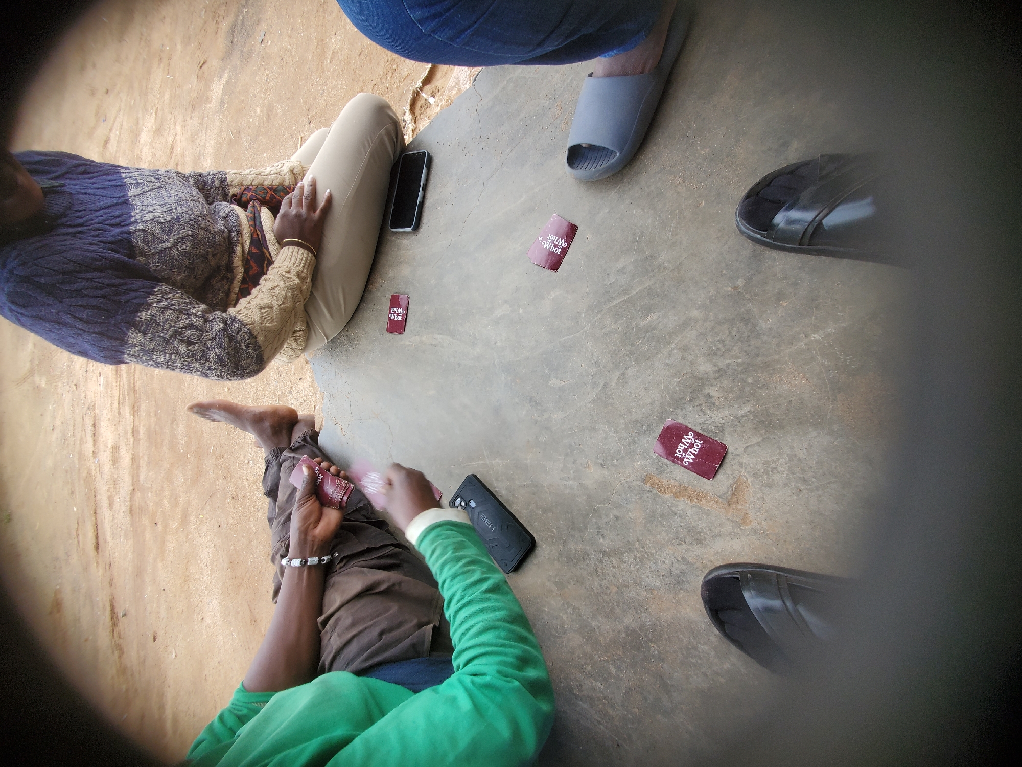 In Bukuru, Nigeria, a group of individuals are engaged in a card game on a concrete surface, likely an outdoor area. The scene is shot from a low angle, with blurred foreground elements obscuring the viewer's perspective.
One person, dressed in a green long-sleeved shirt and brown pants, is seated on the ground with their legs extended. They are holding a stack of red cards in their right hand, and a bracelet adorns their wrist.
Another individual, wearing a multi-colored knitted sweater and light-colored pants, is also seated. They are holding their hands clasped in their lap.
In the middle ground, a black smartphone rests on the concrete, adjacent to a mobile phone case. Several red cards with white text are scattered on the surface, some appearing to be in play. The text on one visible card reads "You Whot".
The environment suggests a dry, outdoor setting with a sandy, dirt ground visible in the background. The overall impression is one of casual social interaction and leisure.
