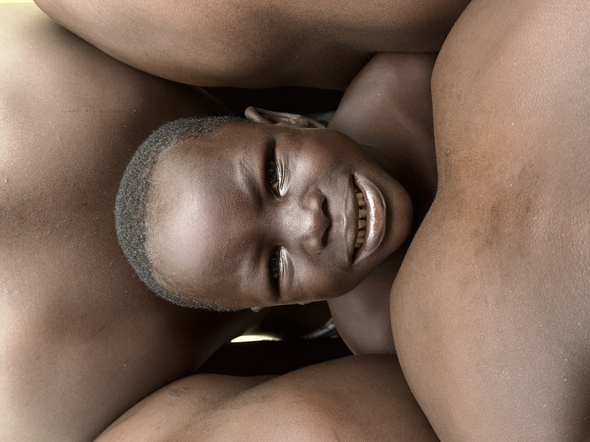 This close-up, eye-level photograph shows a young Black child nestled between the legs of adults. The child's head is tilted upwards, and they are looking directly at the camera with a wide, gap-toothed smile. Their eyes are bright and alert, and their skin is a rich, dark brown. The child's hair is short and appears to be closely cropped. The surrounding legs are also a dark brown, with visible skin texture. The lighting suggests it could be daytime, but the setting is not clearly defined beyond the immediate presence of the people. The overall impression is one of warmth, happiness, and a sense of being embraced or protected. The context of Ado, Nigeria, places the scene within a specific cultural and geographical environment, although no explicit visual cues in the image itself confirm this location. There is no visible text in the image.
