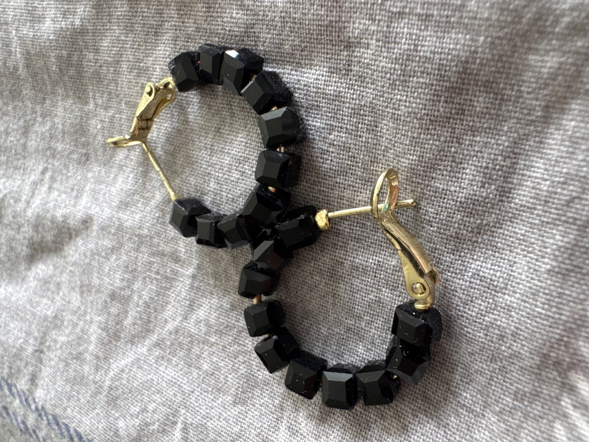 This media file features a close-up, overhead shot of two gold-colored hoop earrings adorned with faceted black beads. The earrings are placed on a textured, gray fabric, likely a textile such as linen or a similar weave, which serves as the background. The lighting suggests it is daytime, with the light source coming from above and slightly to the left, casting subtle shadows. There are no people visible in the image, and the focus is solely on the jewelry. The scene is static, with the earrings resting on the fabric. No specific location cues are evident beyond the implied indoor setting due to the artificial lighting and controlled environment of photographing an object. There is no visible text. The overall mood is neutral and objective, emphasizing the details of the jewelry.