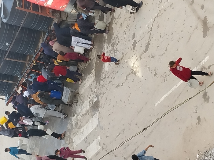 An overhead photograph captures a water distribution scene on a paved street in Khan Yunis, Palestinian Territory. A large red truck with a flatbed loaded with approximately eight prominent black cylindrical water tanks is parked on the right side of the street. A dense crowd of approximately 30-40 individuals, including adults and children, surrounds the truck, actively engaged in filling various plastic containers—such as white, yellow, and blue jerrycans and buckets—from the tanks. Many individuals are bending or reaching towards the truck to access the water.
In the foreground and midground, several children are visible: one child in a red top carries a white jerrycan across the street from right to left; another young child in a red top stands near white road markings; a third small child in red runs in the midground; and a child in a blue shirt carries a white container from left to right. The street itself is concrete or asphalt, marked with two parallel white lines suggesting a pedestrian crossing, and exhibits scattered wet patches. Overhanging electrical cables traverse the upper-left to lower-right portion of the frame. A pile of light-colored rubble is visible in the lower-left corner.